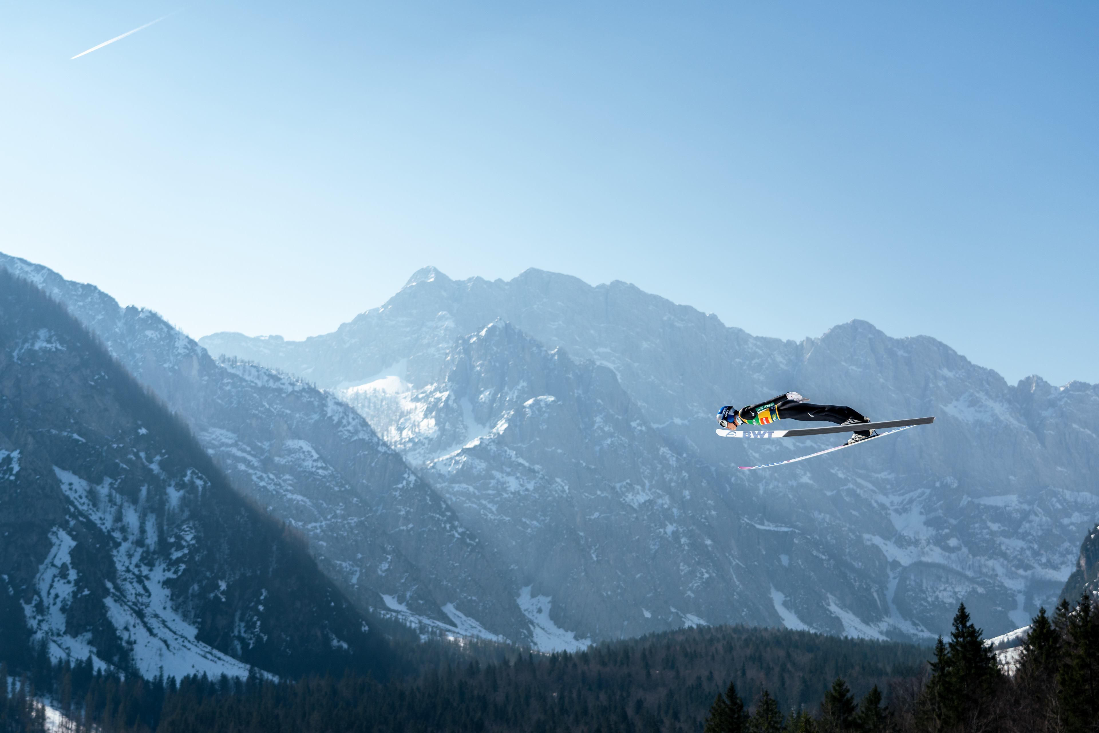 A male Ski Jumper flys dramatically in-front of a rocky, jagged mountain in the late afternoon light. The crisp cold air softens the light and creates a glowing effect! Red Bull Athlete. Slovenia.