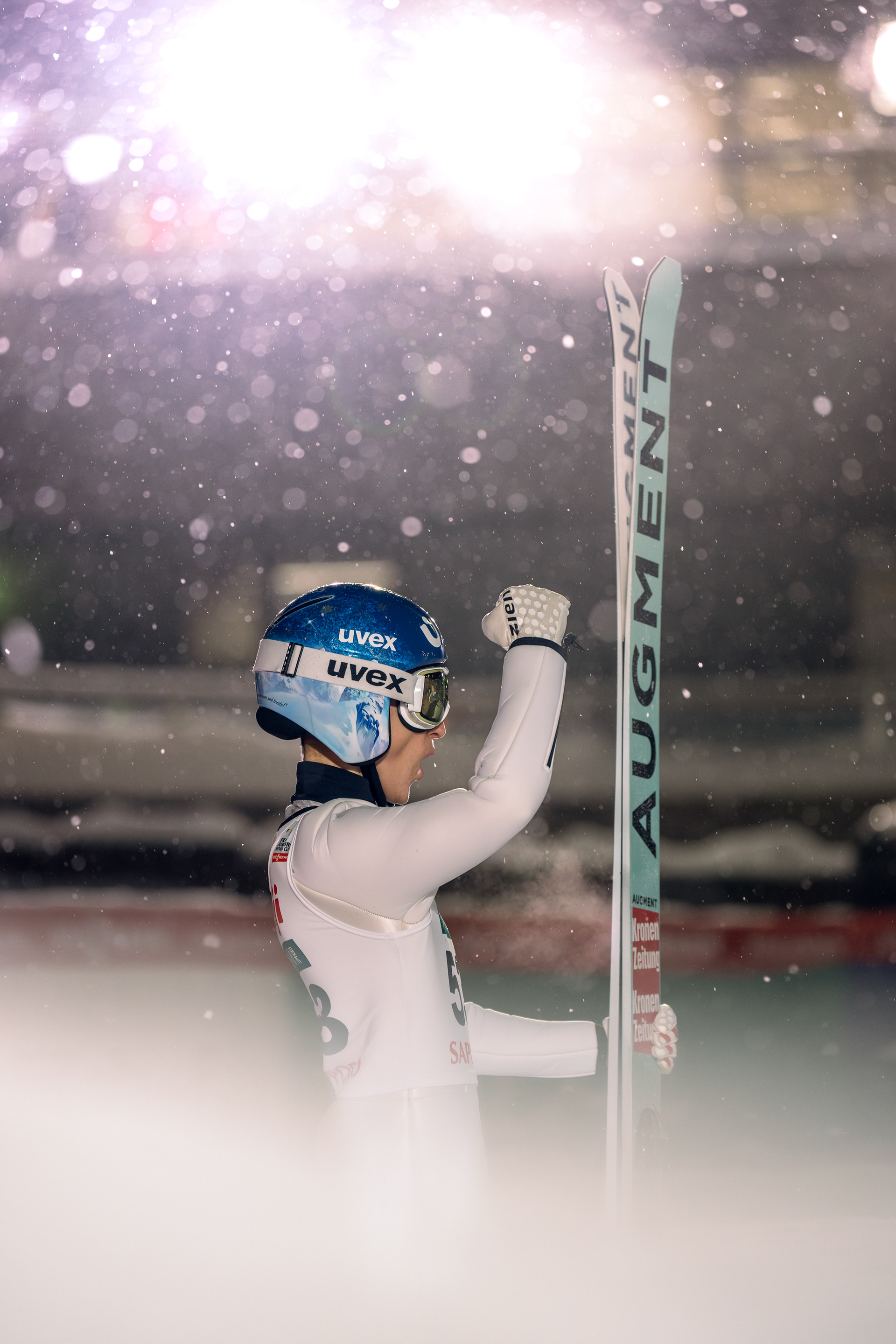 A female Ski jumper stands with her fist pumping to the sky. There is a light in the background illuminating the snow flakes falling. Action Sports Photographer.