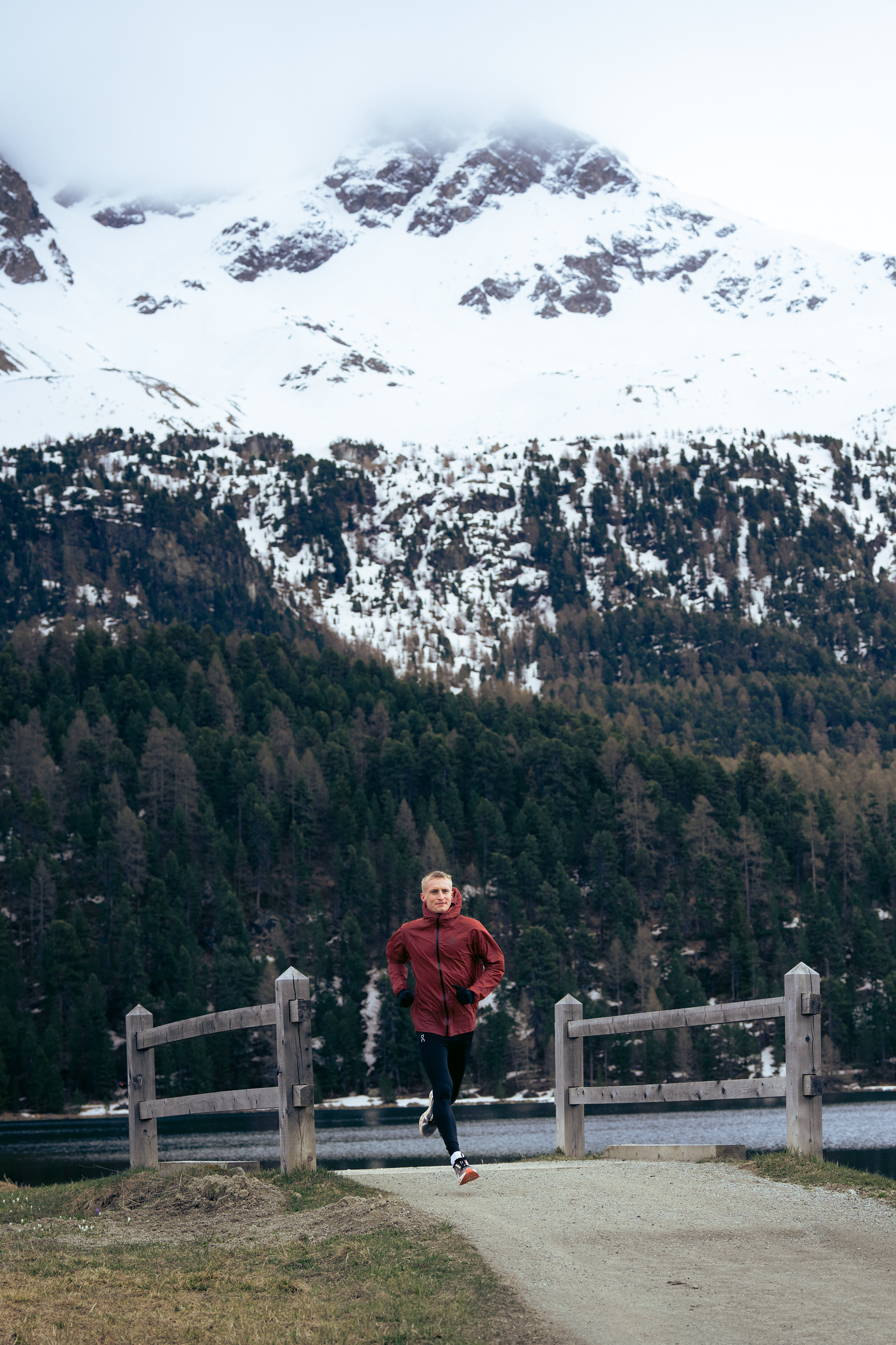 A male runner strides across a small wooden bridge infant of a lake and Large snow capped Mountain.
