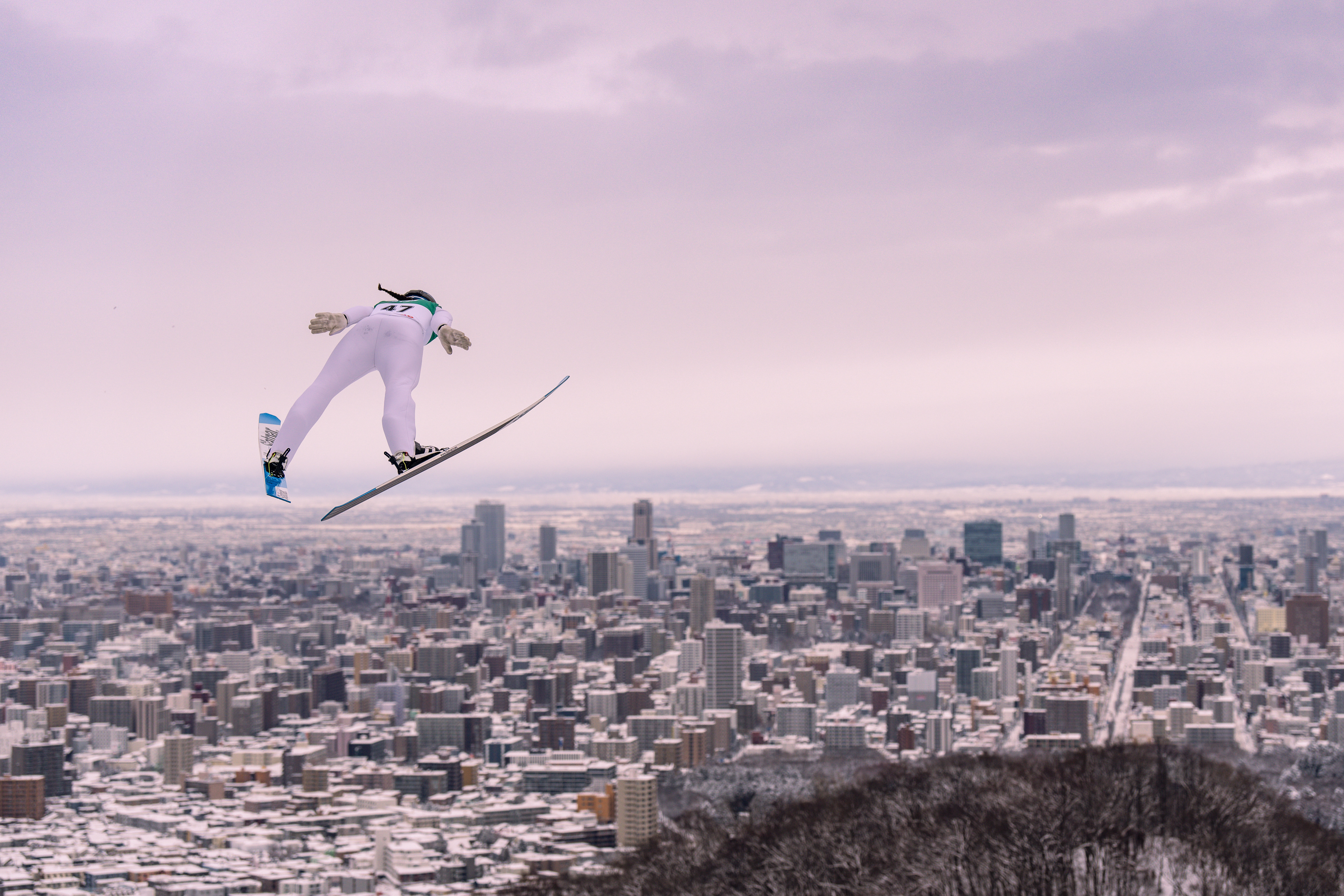 A female ski Jumper, Alex Loutitt, Jumps above the city scape of Sapporo Japan. The sky is overcast and a soft purple. Canadian Ski Jumper. High Rise cityscape of Sapporo, Japan.