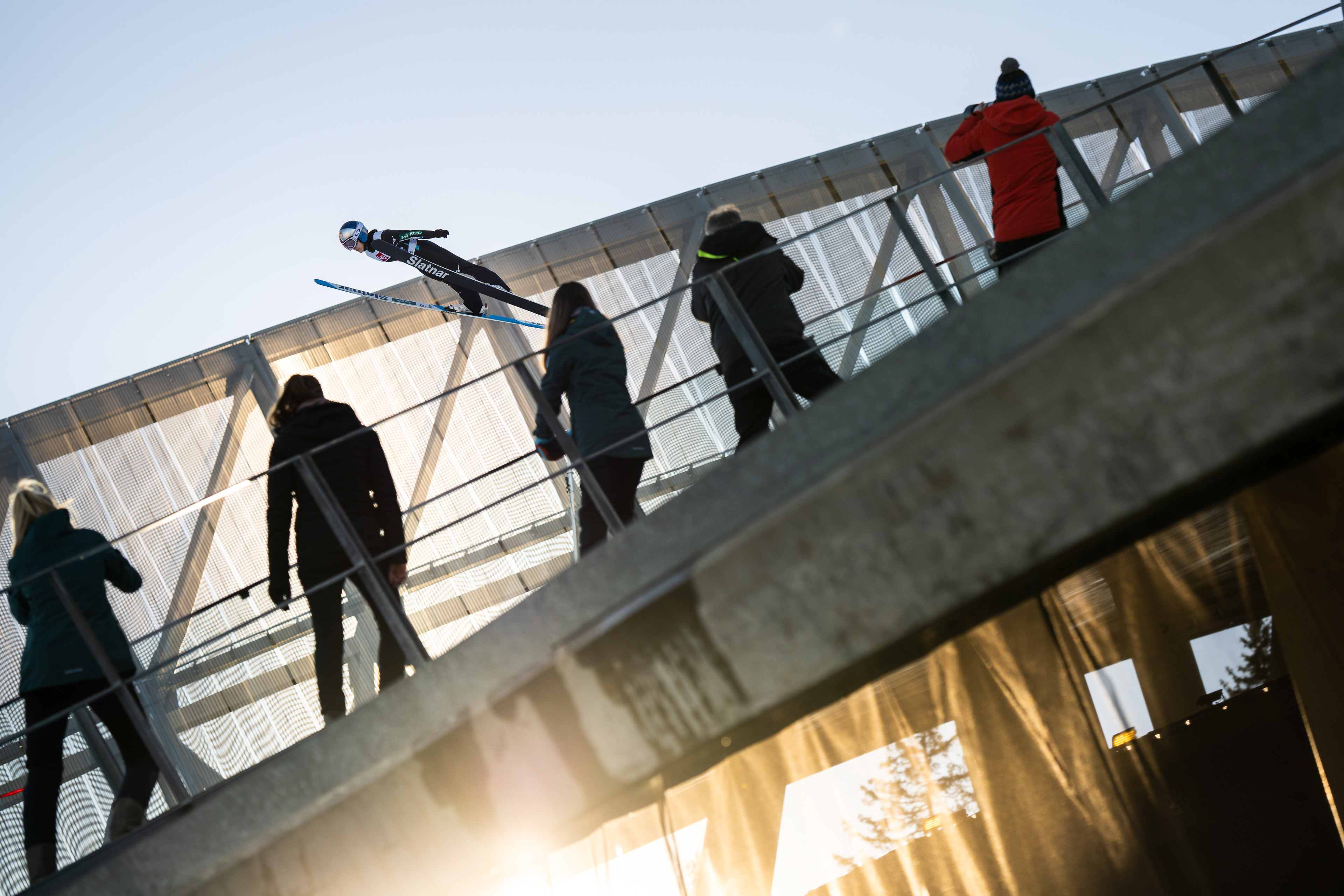 Sara Takanashi, A Japanese female Ski Jumper, jumps under the focus of multiple photographers and videographers at Holmenkollen. The light is golden, and the sun is setting in the background. Oslo, Norway. Red Bull Athlete. Unique perspective. Professional Sports Photographer.