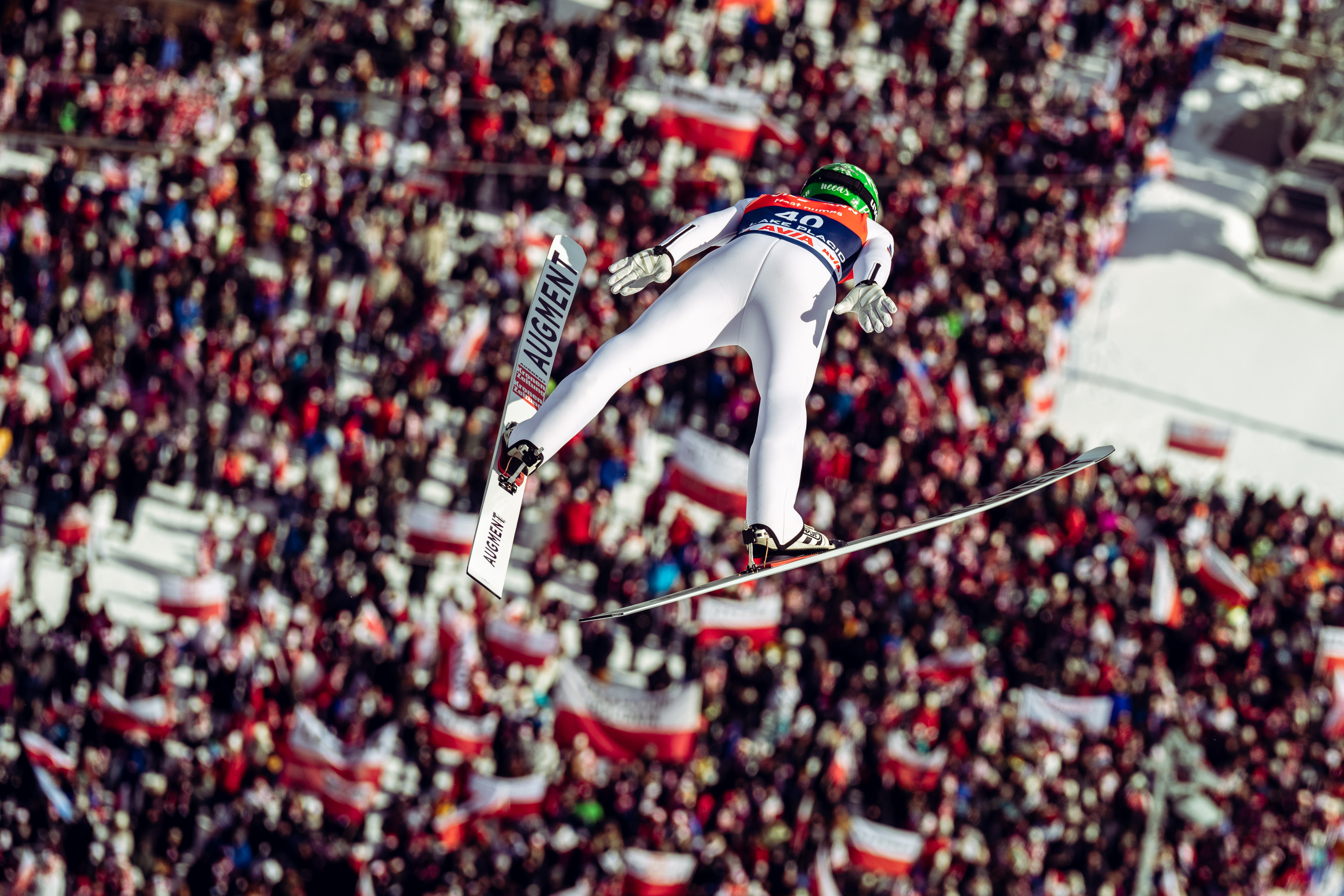 A male Ski Jumper in wite flys above a crowd of thousands of people waving flags and banners.
