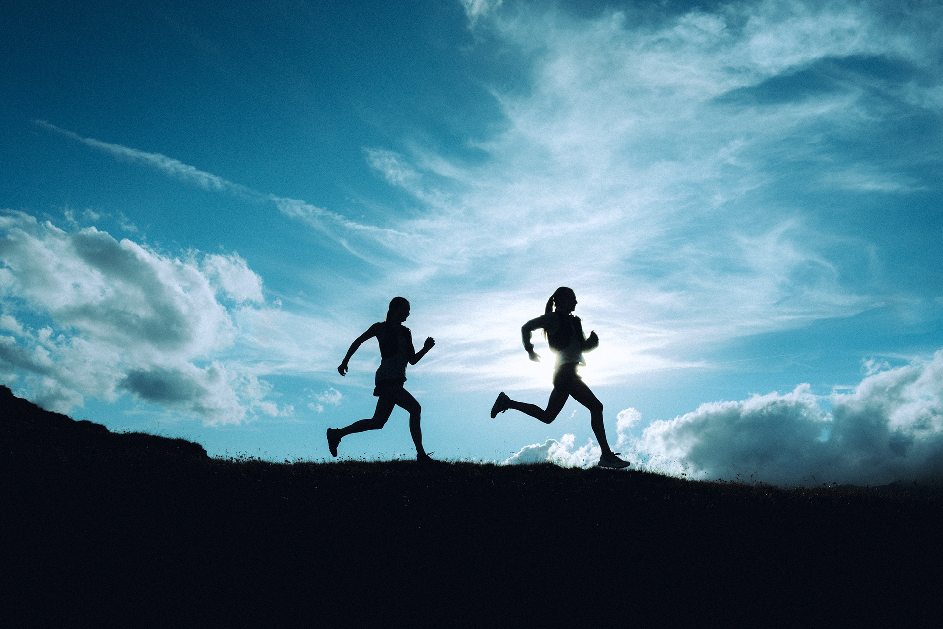 Two female silhouettes running along a ridge in front of the sun and a Partially cloudy sky