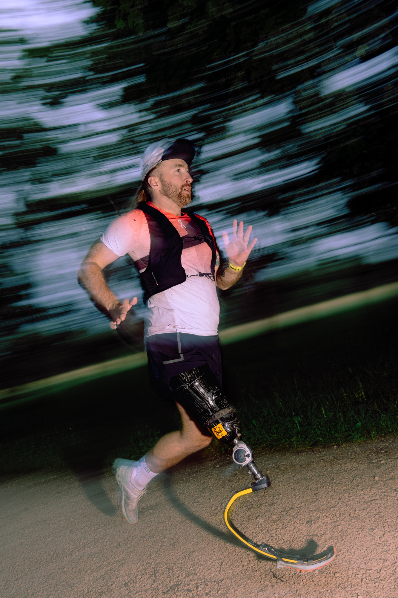 A male runner, photographed with motion, moving through some trees on a dirt path. The Runner has one running blade and one leg.