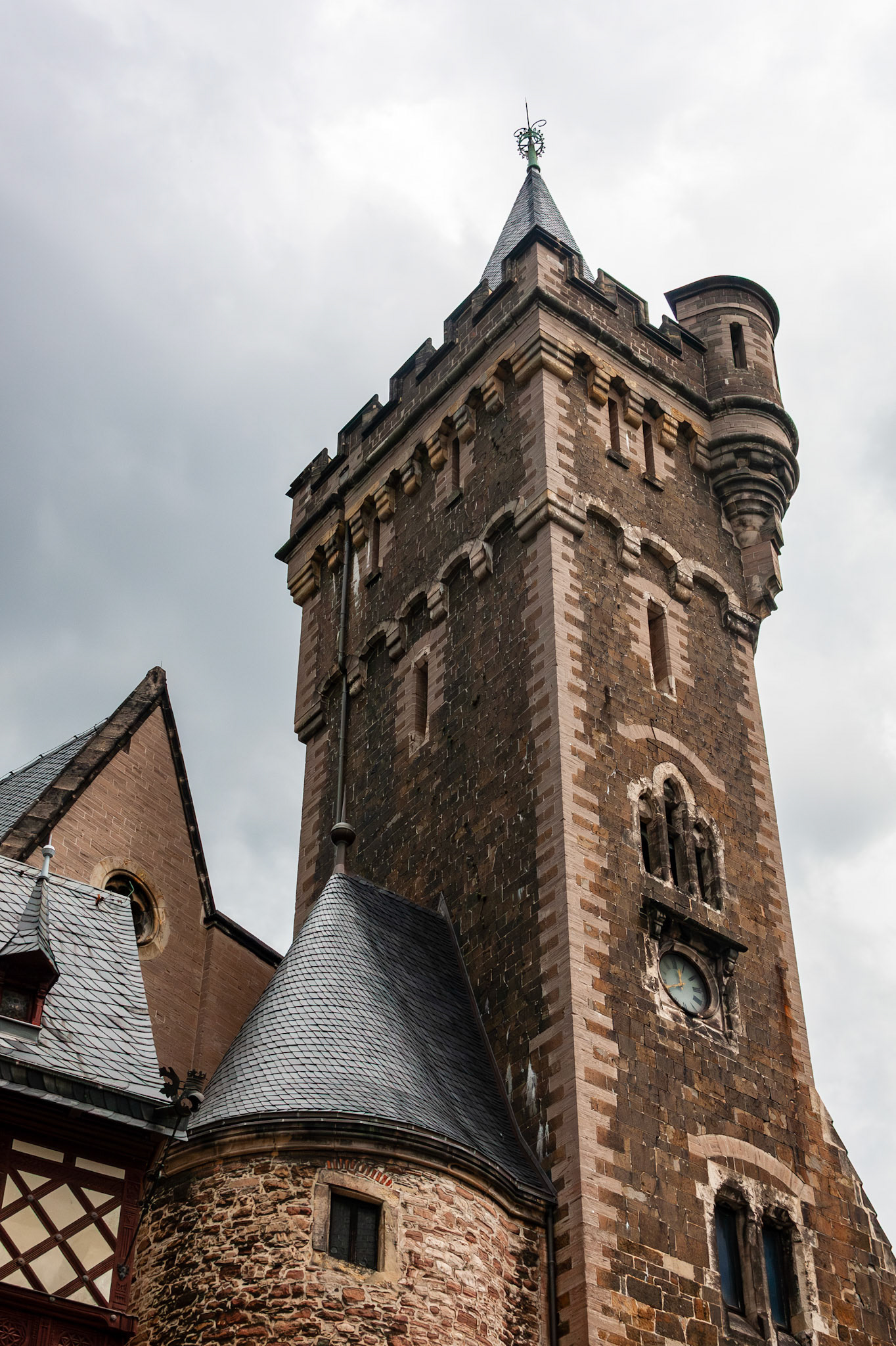 A dramatic low-angle shot of a tall, medieval stone tower at Wernigerode Castle, featuring a clock face, arched windows, and a pointed spire under a moody grey sky in Germany.