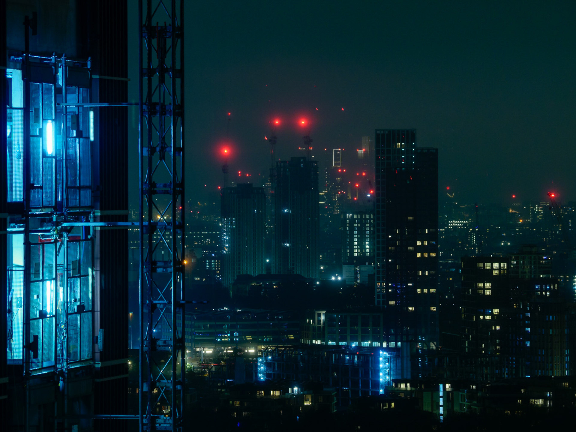 A moody, futuristic night view of the London cityscape, featuring a neon-blue illuminated construction elevator in the foreground against a backdrop of dark skyscrapers and glowing red aviation lights.