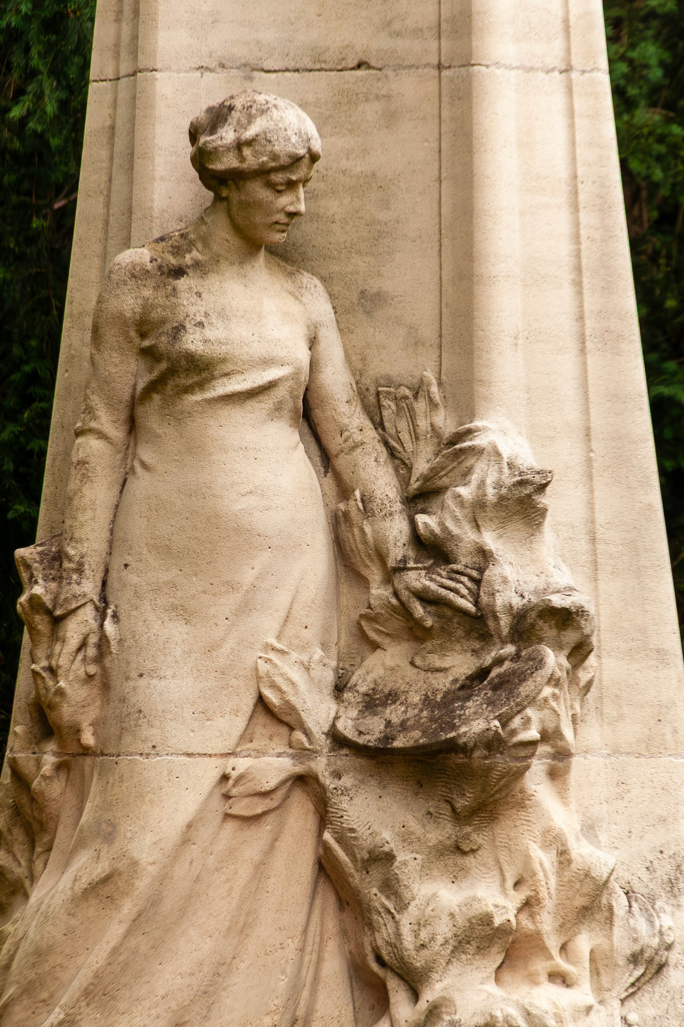 A graceful stone relief sculpture of a woman in classical attire, carved into a monument pillar in Nancy, France. The figure is depicted in a pensive pose, surrounded by delicate floral and leaf motifs characteristic of the Art Nouveau style.