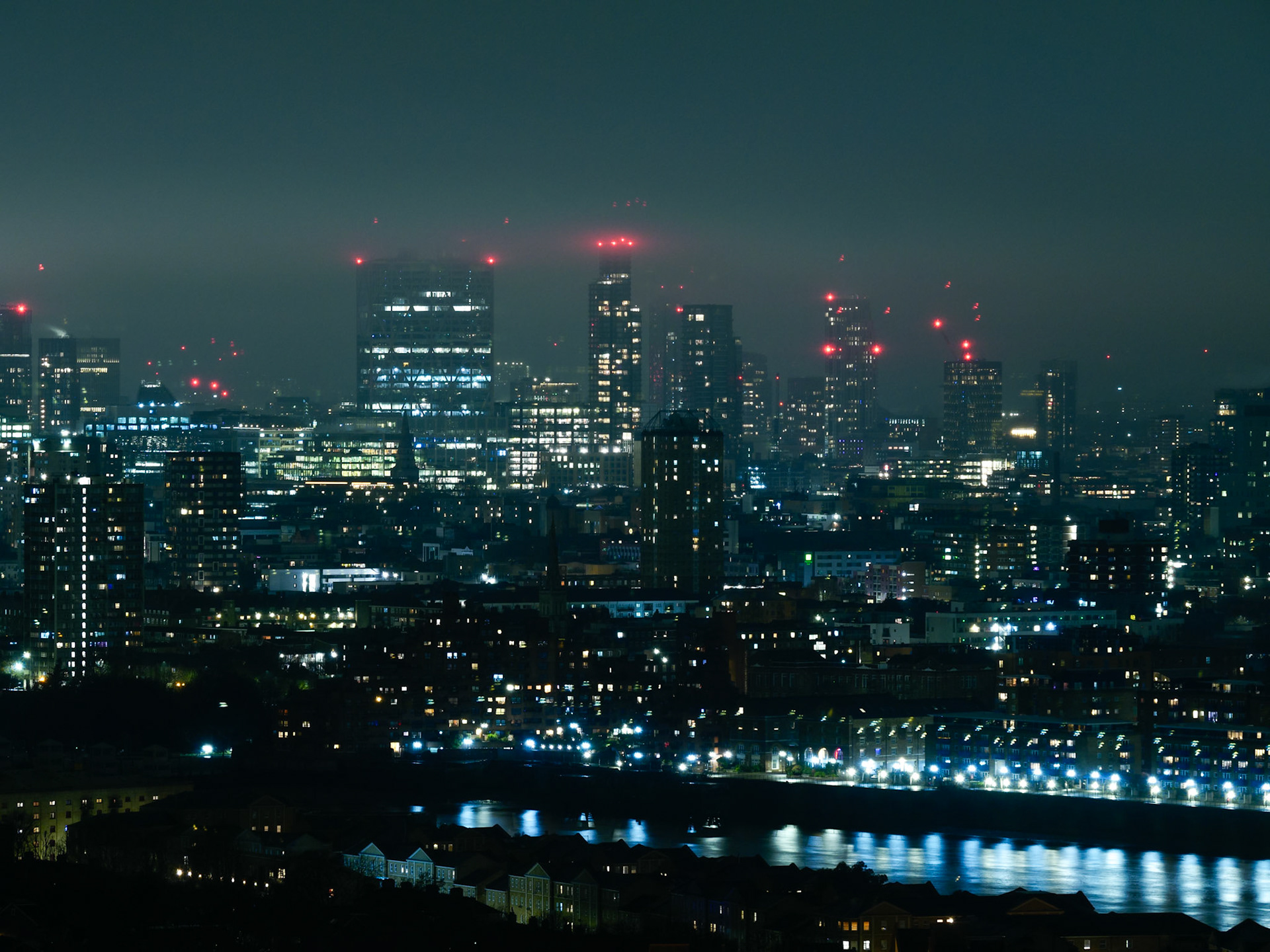 A wide, atmospheric night view of the London skyline featuring glowing skyscrapers and residential blocks reflected in the River Thames, captured with a moody cyberpunk aesthetic under a hazy, dark sky.