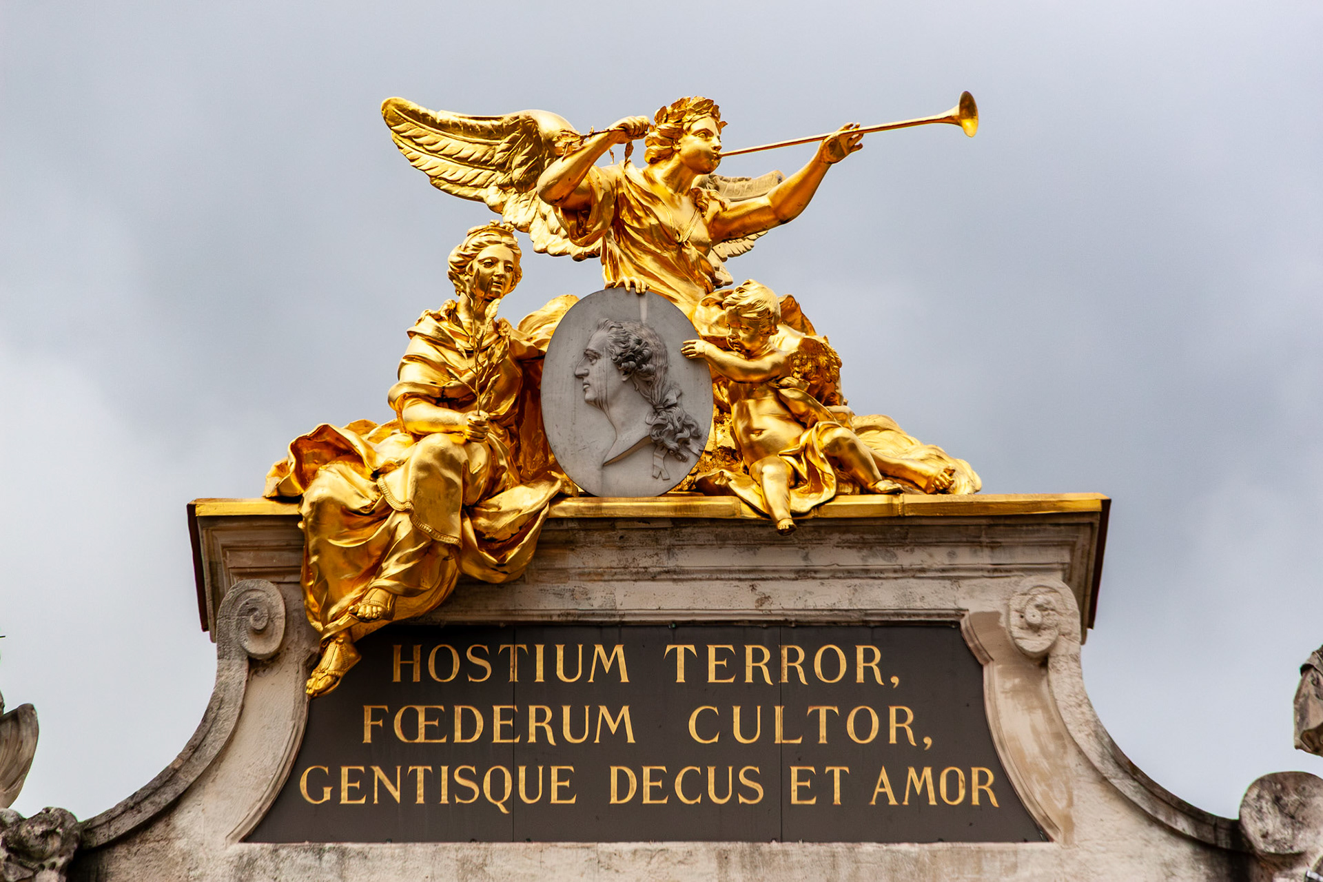 A stunning low-angle shot of a gold-leafed neoclassical sculpture group atop an archway in Place Stanislas, Nancy, France. The group features a winged figure with a trumpet and seated allegorical figures surrounding a central medallion, positioned above a Latin inscription.