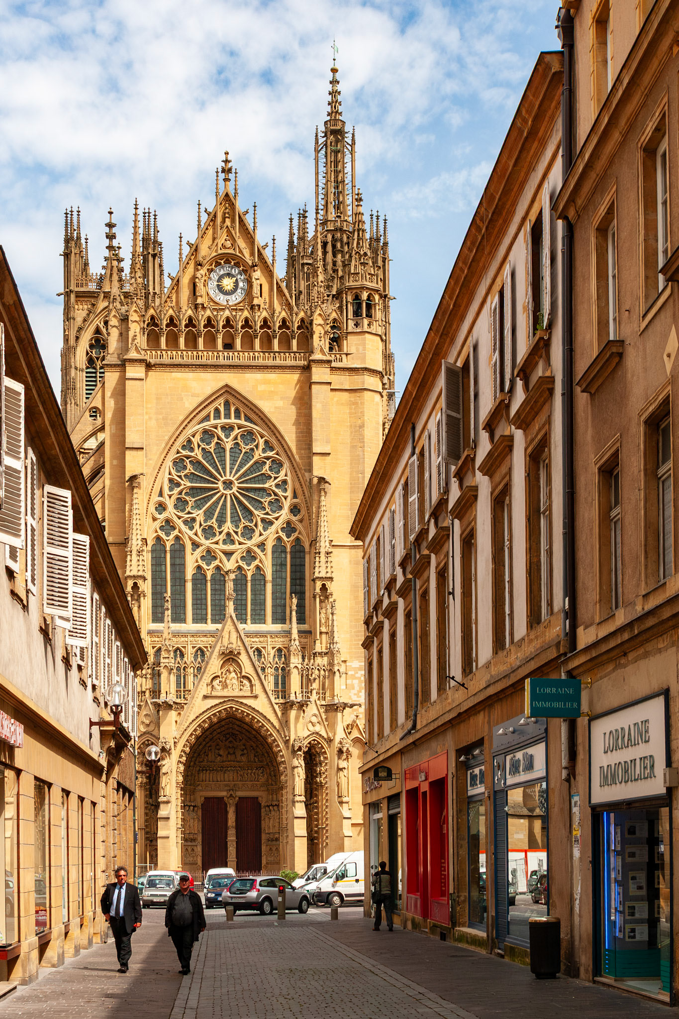 A stunning vertical view of the western facade of Saint-Stephen Cathedral (Cathédrale Saint Étienne) in Metz, France, featuring its massive rose window and intricate Gothic carvings, framed by the traditional buildings of Rue des Jardins.