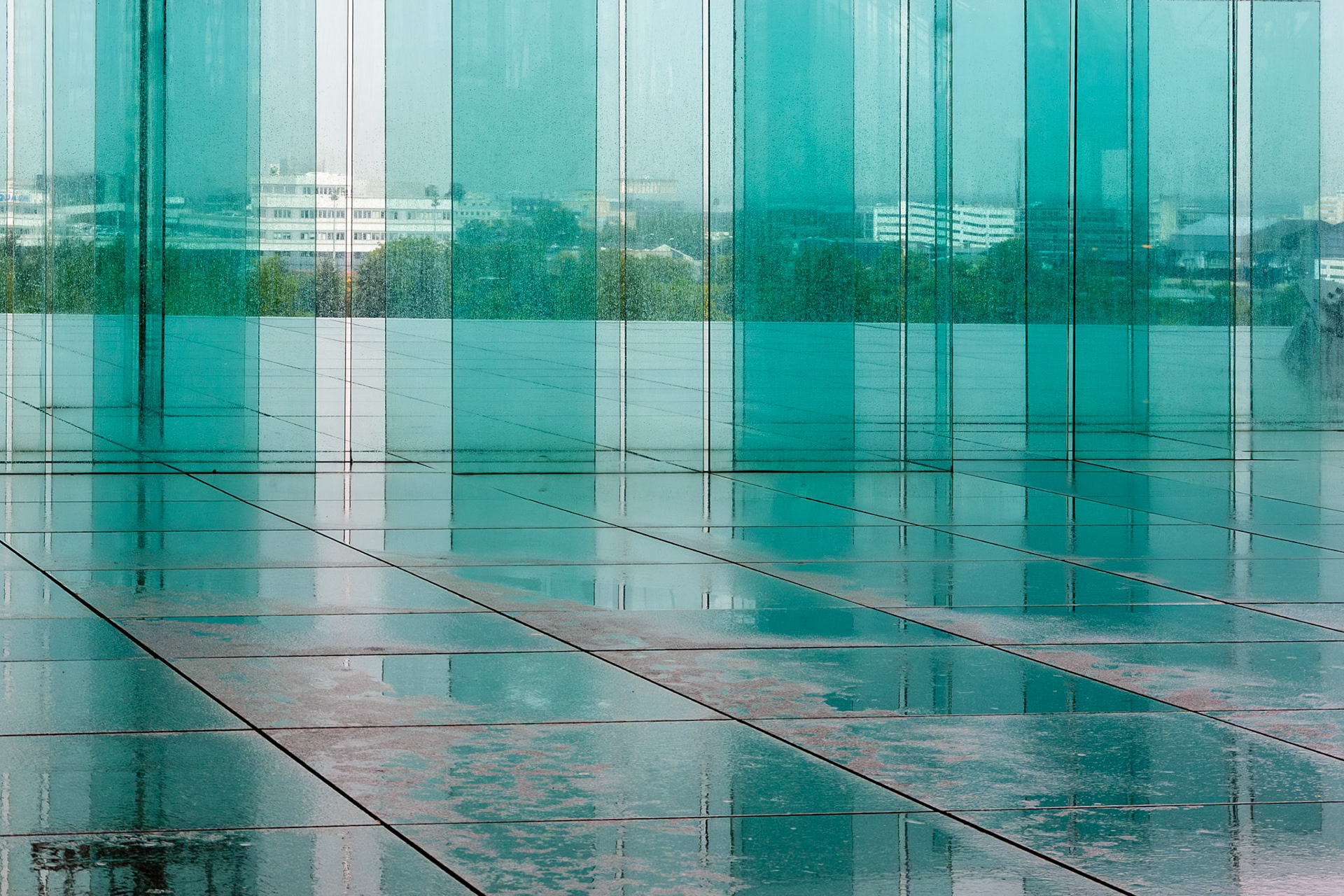 A conceptual shot of contemporary architecture featuring vertical teal glass partitions. The foreground shows a polished, wet stone surface reflecting the turquoise glass and the overcast sky, creating a minimalist and layered geometric composition.