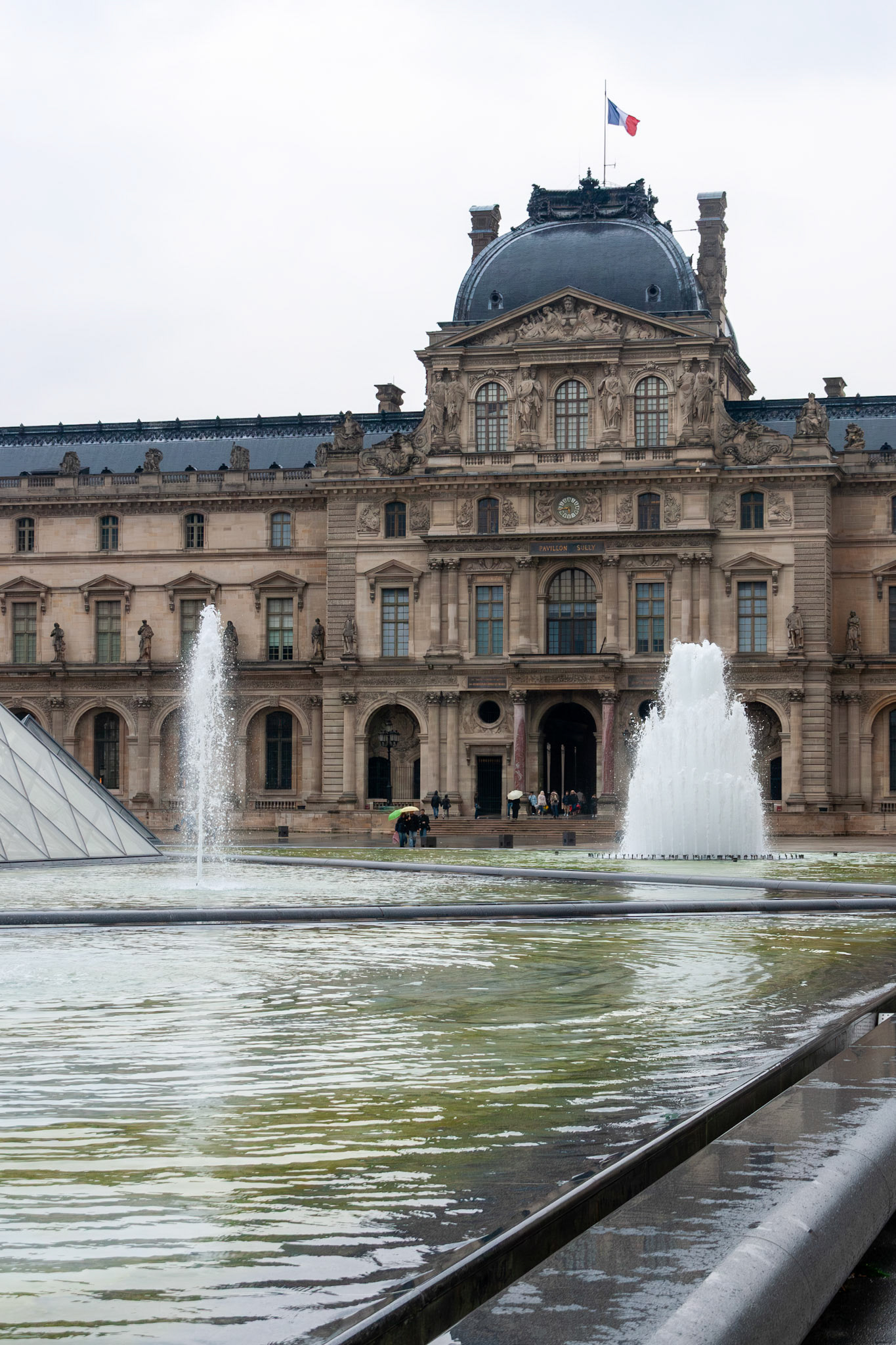 A scenic view of a majestic historic palace in France, featuring ornate Renaissance-style architecture, a fluttering national flag, and active water fountains in a large courtyard under an overcast sky.