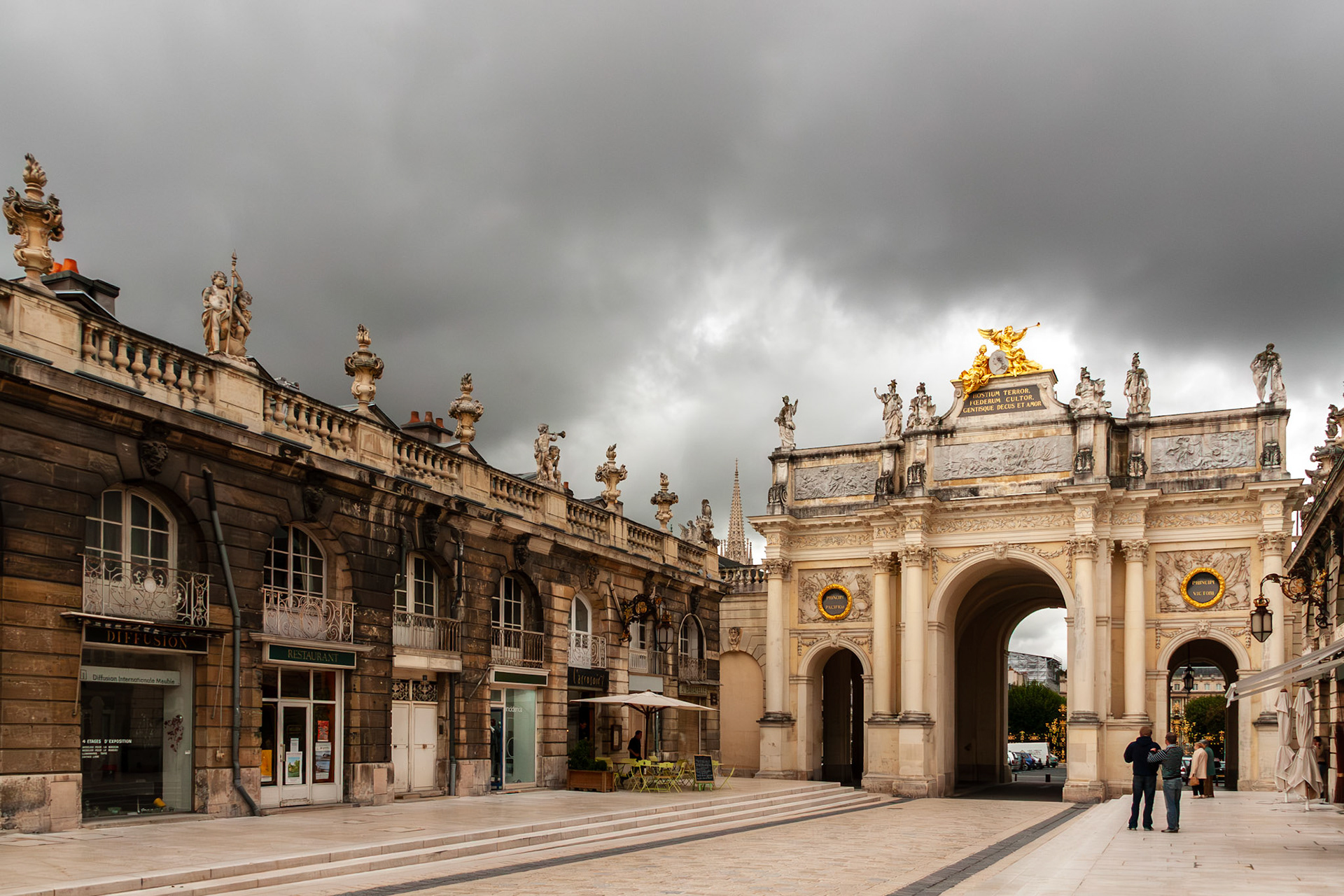A wide perspective of the Arc Héré (Porte Héré) triumphal arch under a moody, overcast sky, flanked by historic neoclassical buildings and restaurants in the UNESCO World Heritage site of Nancy, France.