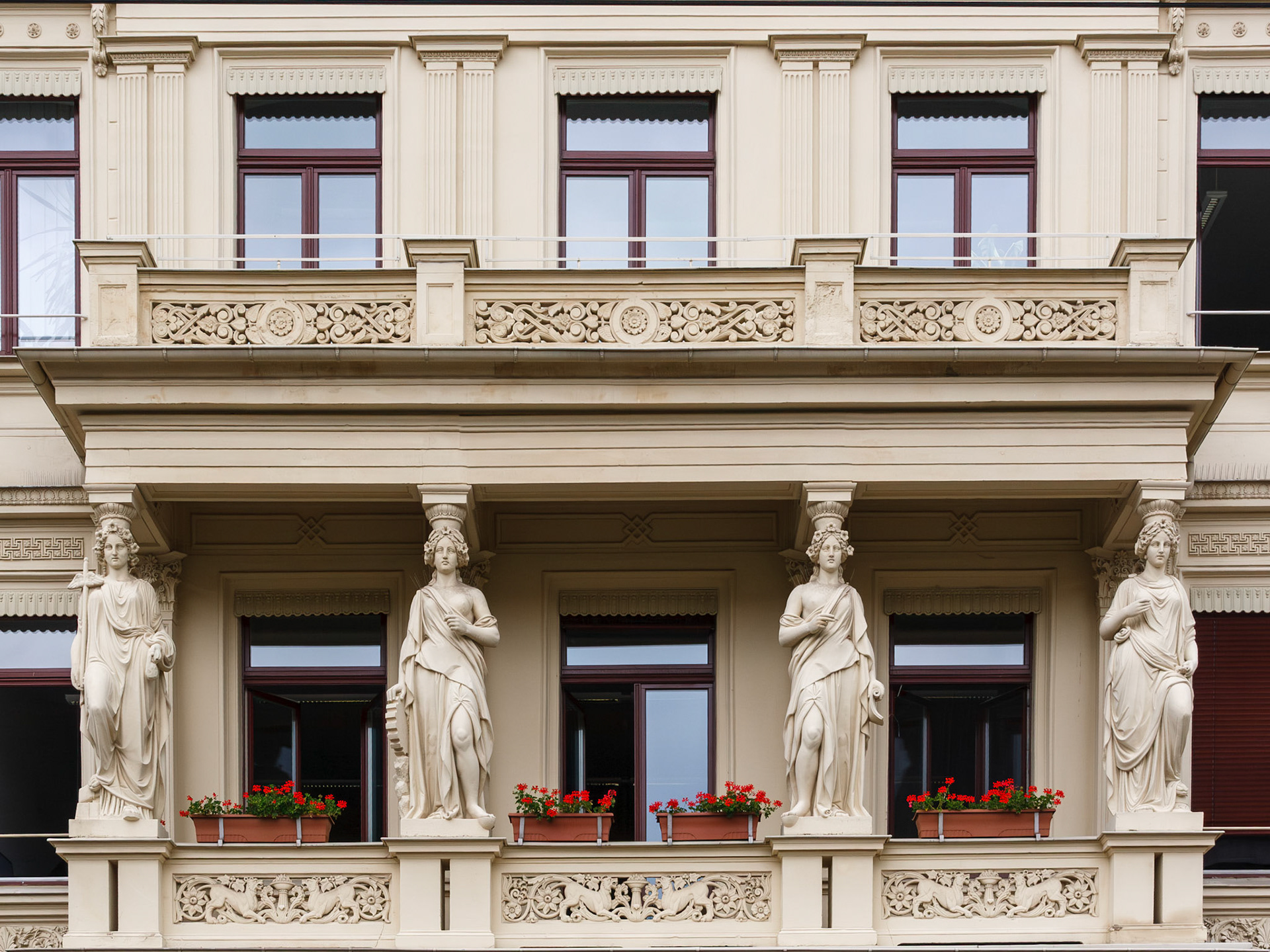 An elegant architectural close-up showcasing four detailed caryatid statues supporting a grand balcony in Görlitz, Germany. The ornate beige facade is decorated with classical carvings, symmetry, and vibrant red flowers lining the window boxes.