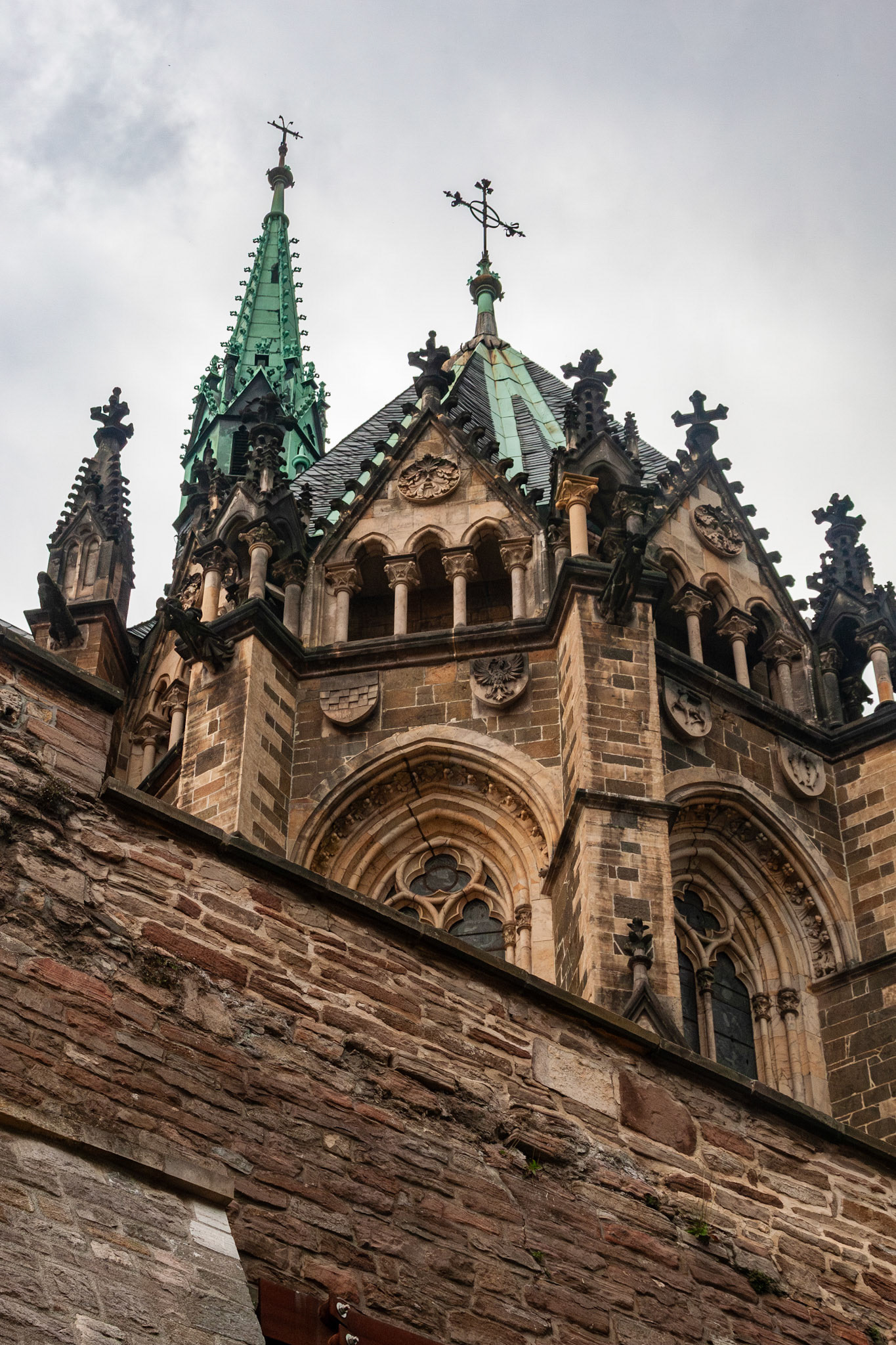 An upward perspective of the intricate Gothic Revival architecture of the Schlosskirche (Castle Church) at Wernigerode Castle, featuring ornate stone carvings, pointed arches, and a prominent green copper spire under an overcast sky in Germany.