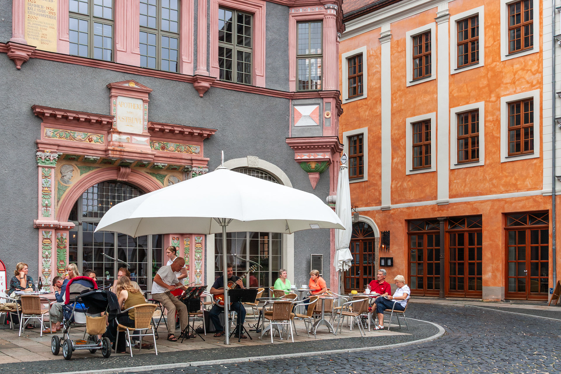 Patrons enjoy refreshments at an outdoor terrace while musicians perform under a large white umbrella, set against the backdrop of vibrant, Renaissance-style architecture in the historic town of Görlitz, Germany.