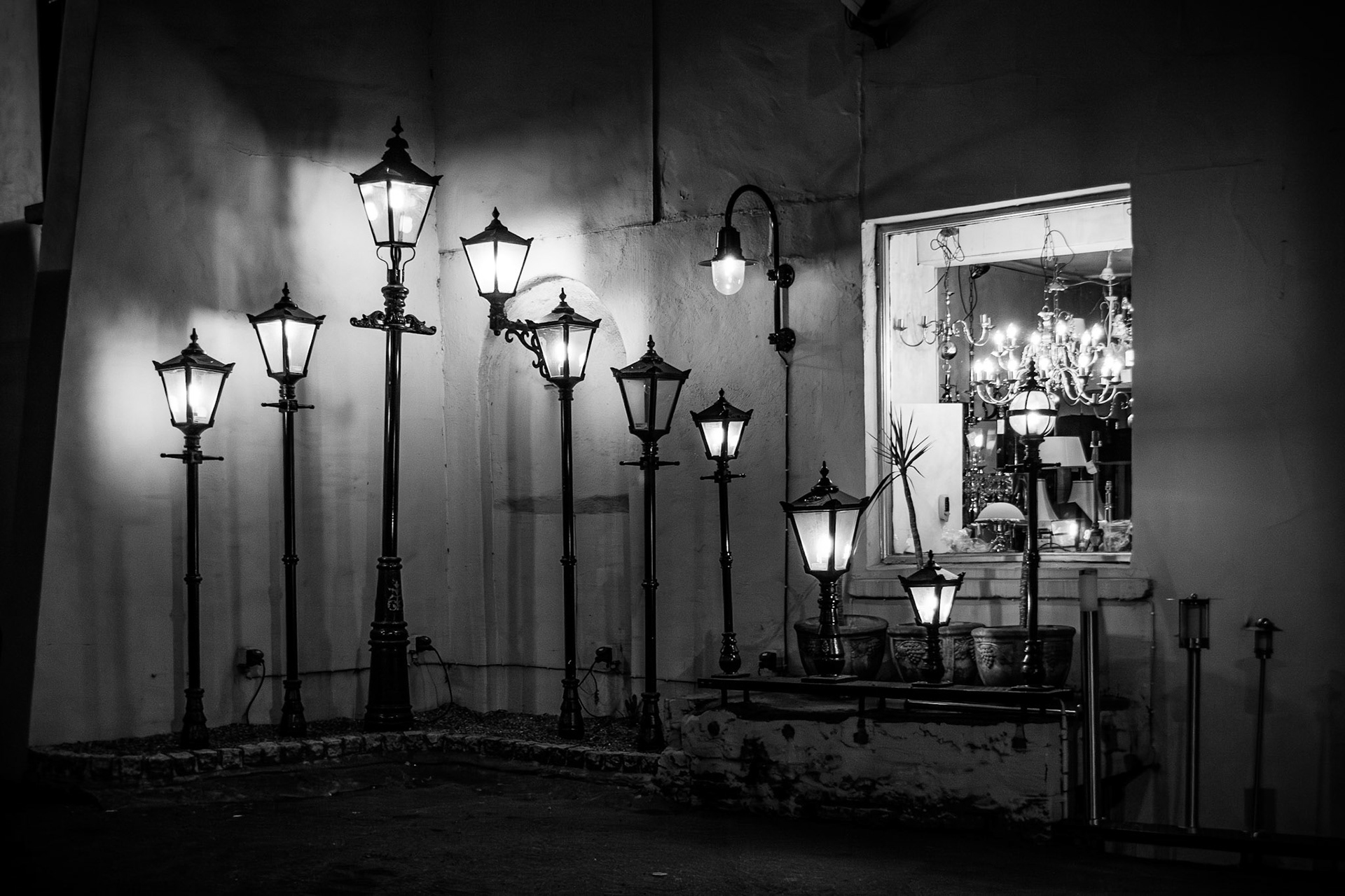 A dramatic black and white night study of various classical street lamp designs illuminated against a dark wall, featuring intricate metalwork and a glowing shop window display of chandeliers.