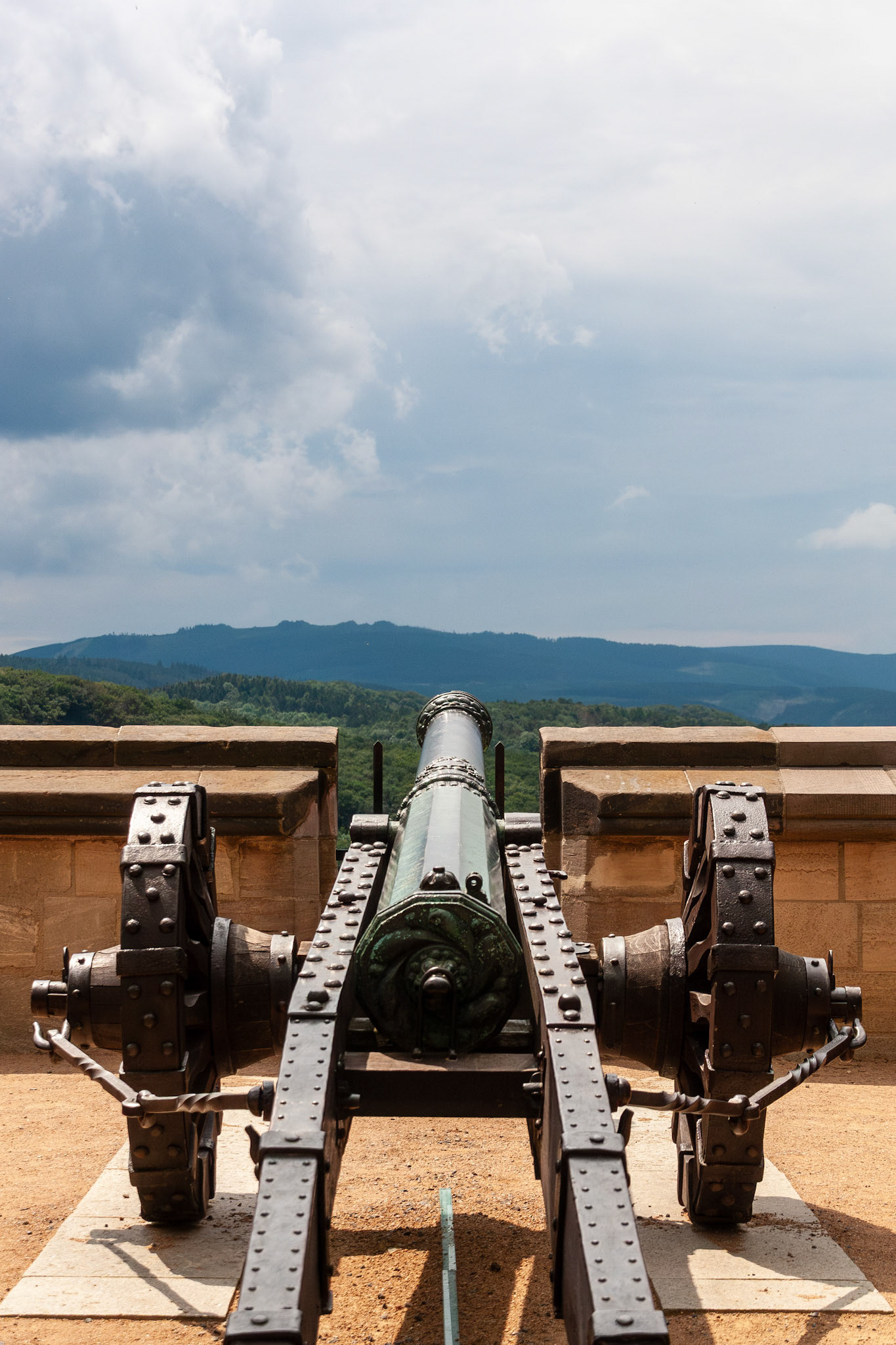 A detailed rear view of an ancient bronze and iron cannon positioned on the stone ramparts of Wernigerode Castle, pointing toward the rolling green hills of the Harz region in Germany.