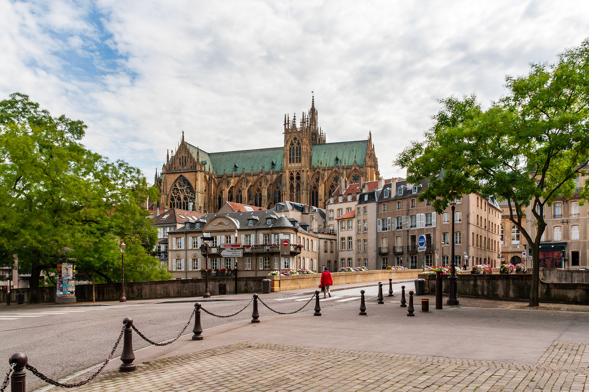 A wide cityscape of Metz, France, featuring the massive Gothic architecture of Saint Stephen Cathedral towering over a row of historic townhouses, a stone bridge with iron bollards, and lush green trees under a cloudy sky.