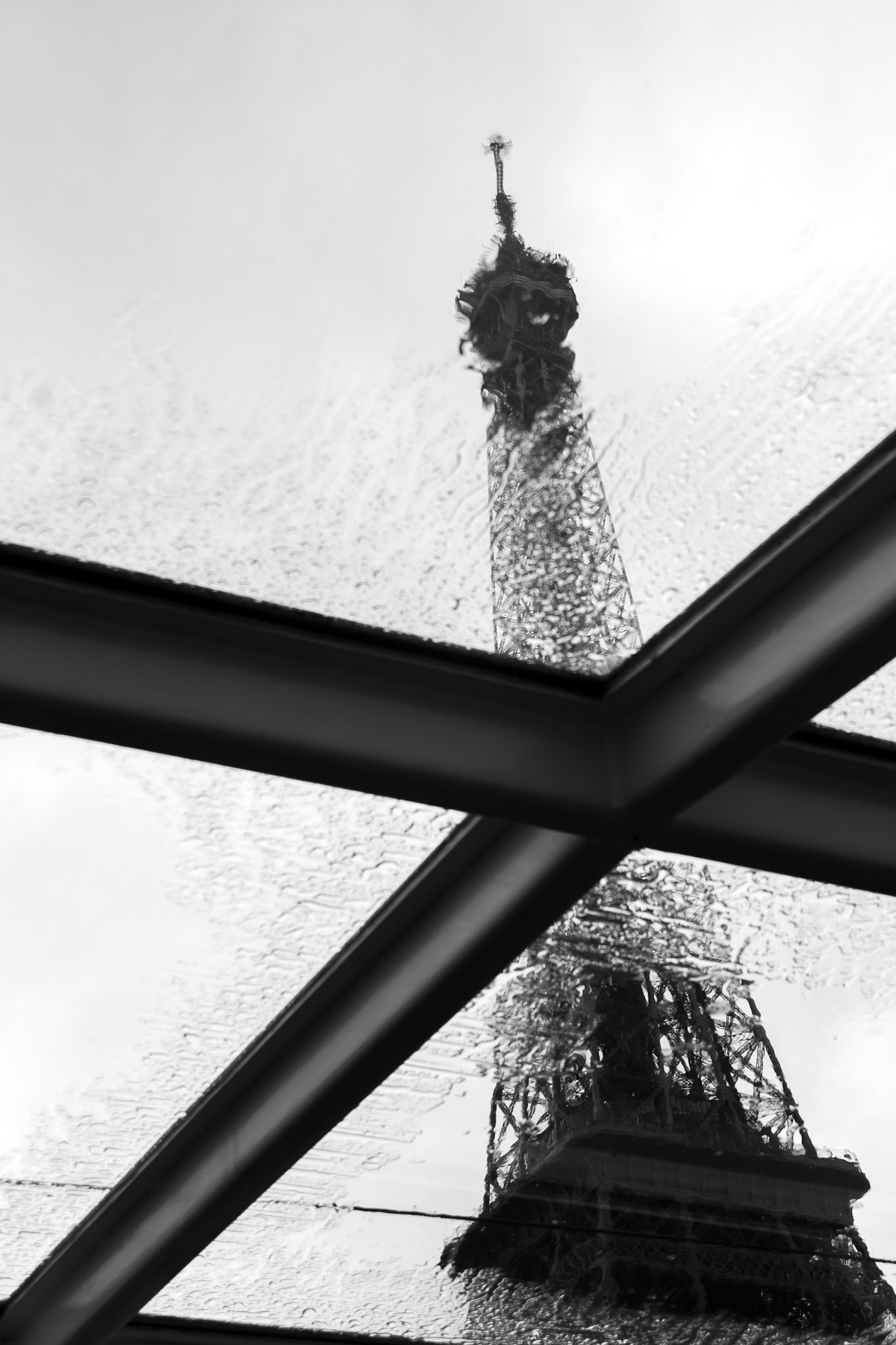 An artistic black and white shot showing the reflection of the Eiffel Tower on a wet glass surface during a rainy day in Paris. The architectural geometry of the window frames intersects the distorted, shimmering reflection of the iconic landmark, creating an abstract and moody atmosphere.