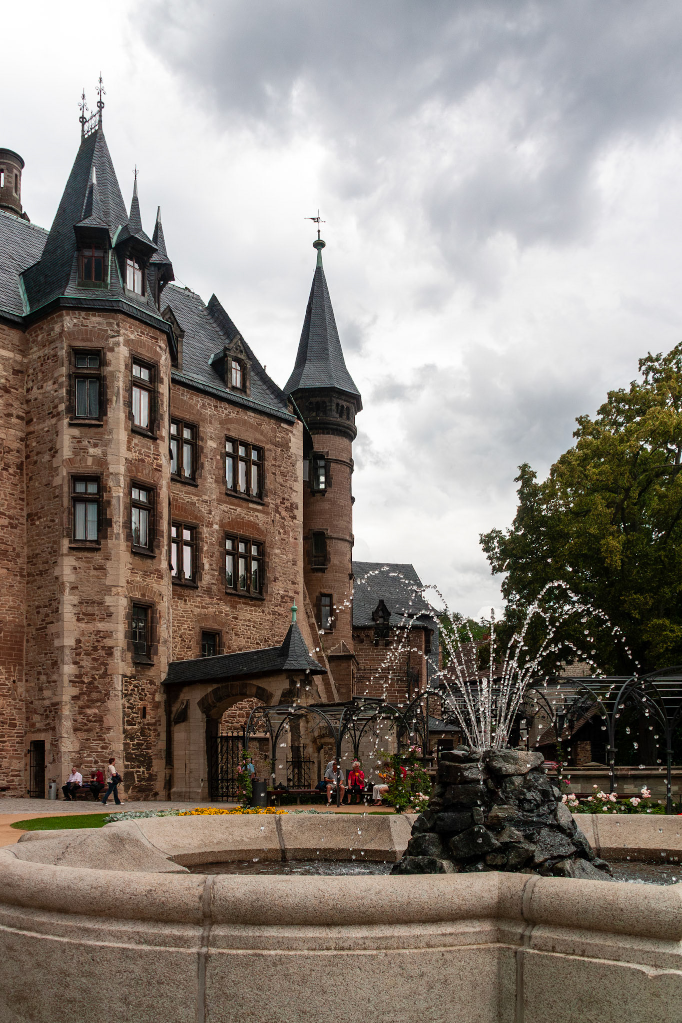 A view of the ornate stone facade and pointed turrets of Wernigerode Castle in Germany, featuring a central tiered fountain in the foreground and a moody, overcast sky.