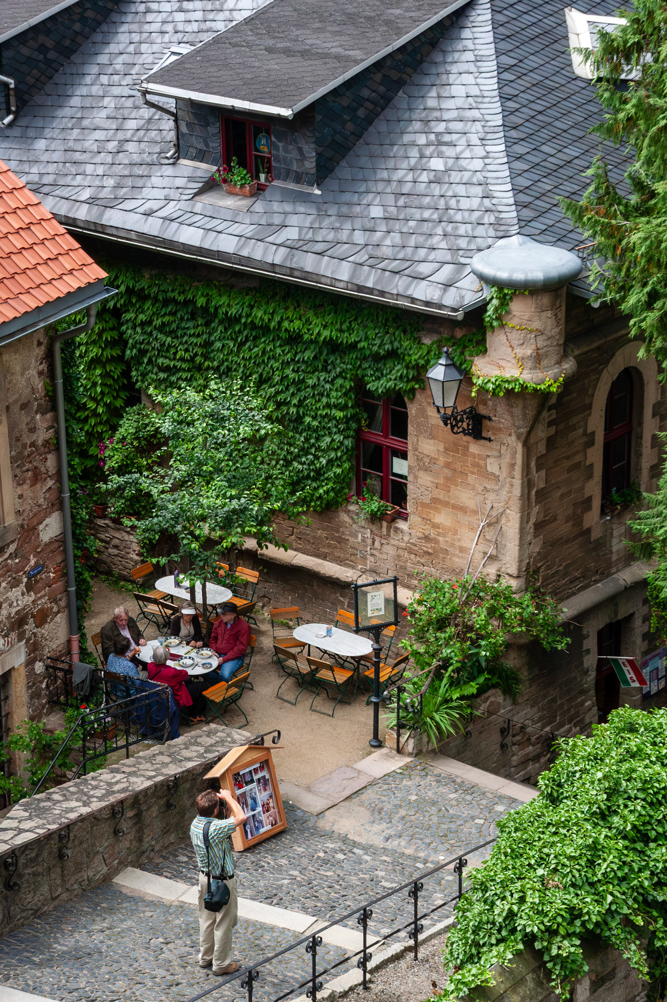 An elevated perspective of a charming, ivy-clad courtyard café at Wernigerode Castle in Germany, featuring patrons dining at white tables surrounded by historic stone architecture and lush greenery.
