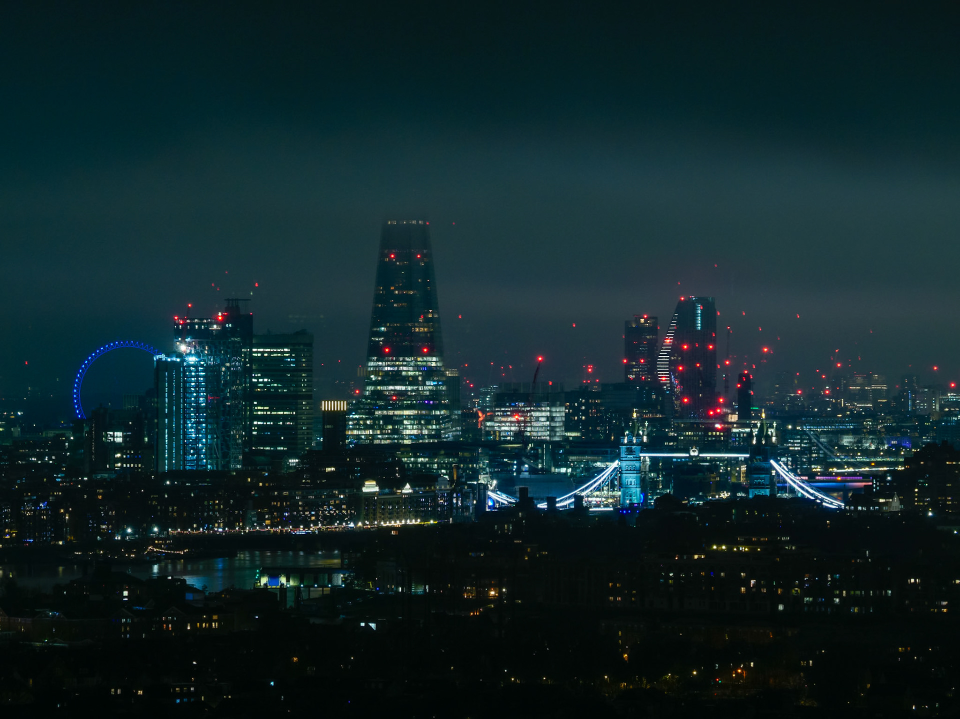 A panoramic night view of the London skyline featuring iconic landmarks including The Shard, Tower Bridge, and the London Eye, all illuminated with a moody, futuristic cyberpunk aesthetic under a dark misty sky.
