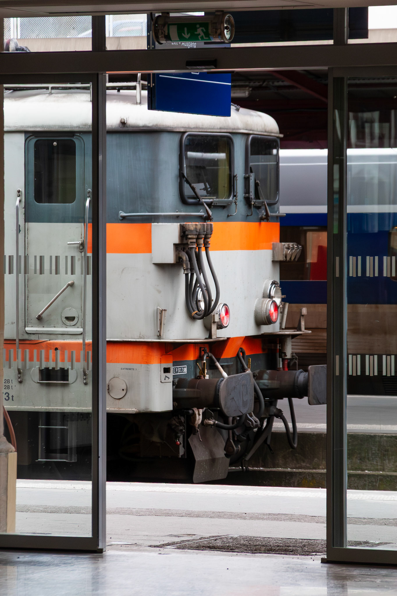 A view through glass station doors of a grey and orange BB 16500-style electric locomotive stationary at a platform in Nancy, France.