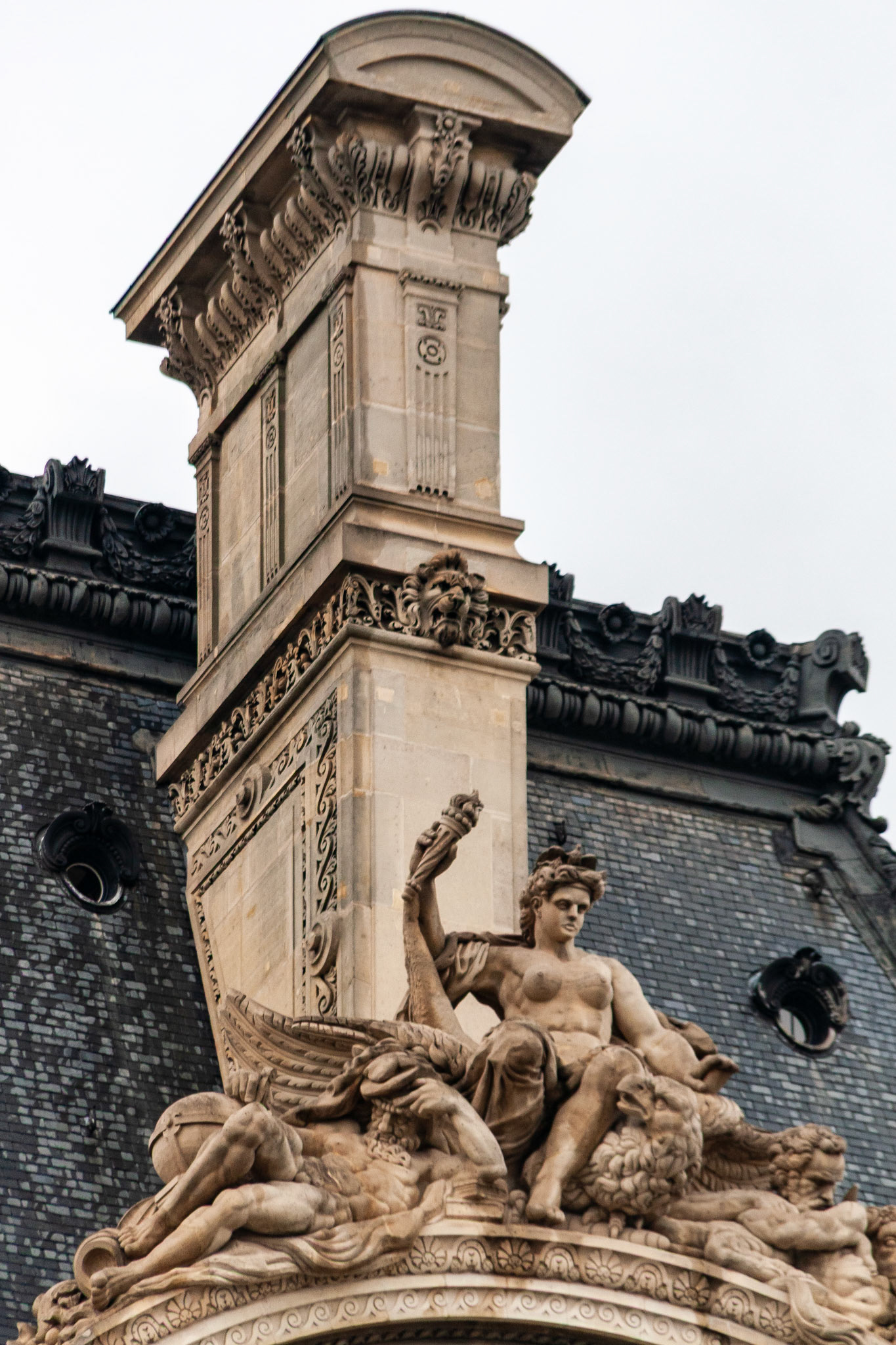 An intricate limestone sculpture and ornate chimney detail on the roof of a classic Neo-Renaissance building in Paris, France. The image features a central female figure holding a torch, flanked by mythical creatures and elaborate stonework against a overcast sky.