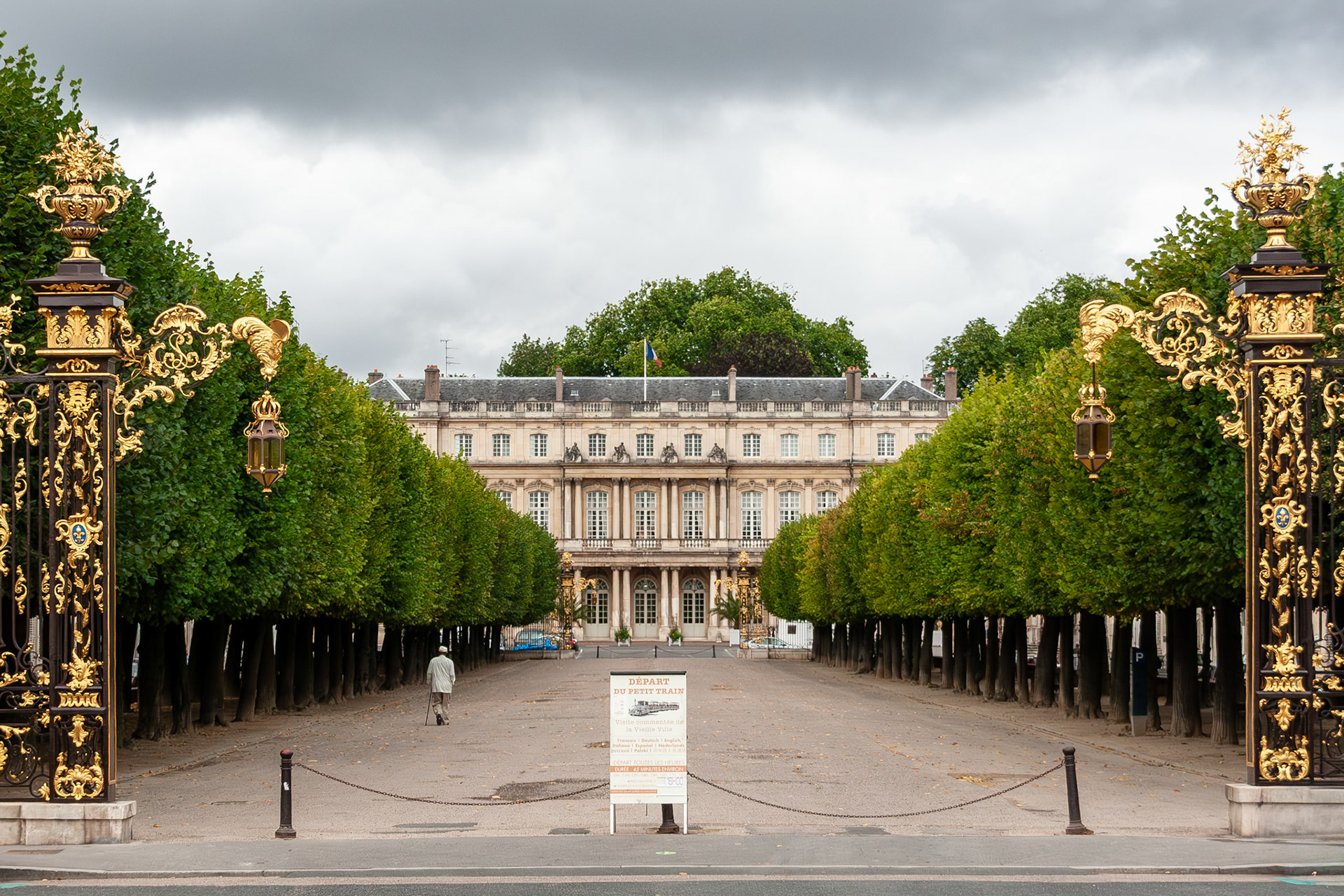 A scenic view looking down a tree-lined path toward the neoclassical Palais du Gouvernement in Nancy, France, framed by the iconic black and gold wrought-iron gates of the Place de la Carrière.
