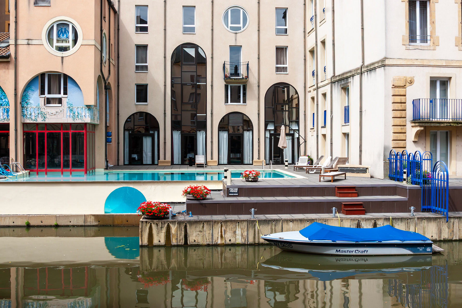 A view of contemporary waterfront architecture in Metz, France, featuring a residential building with a private terrace and swimming pool. A blue MasterCraft boat is docked at the edge of the Moselle River, which reflects the clean lines and large arched windows of the facade.