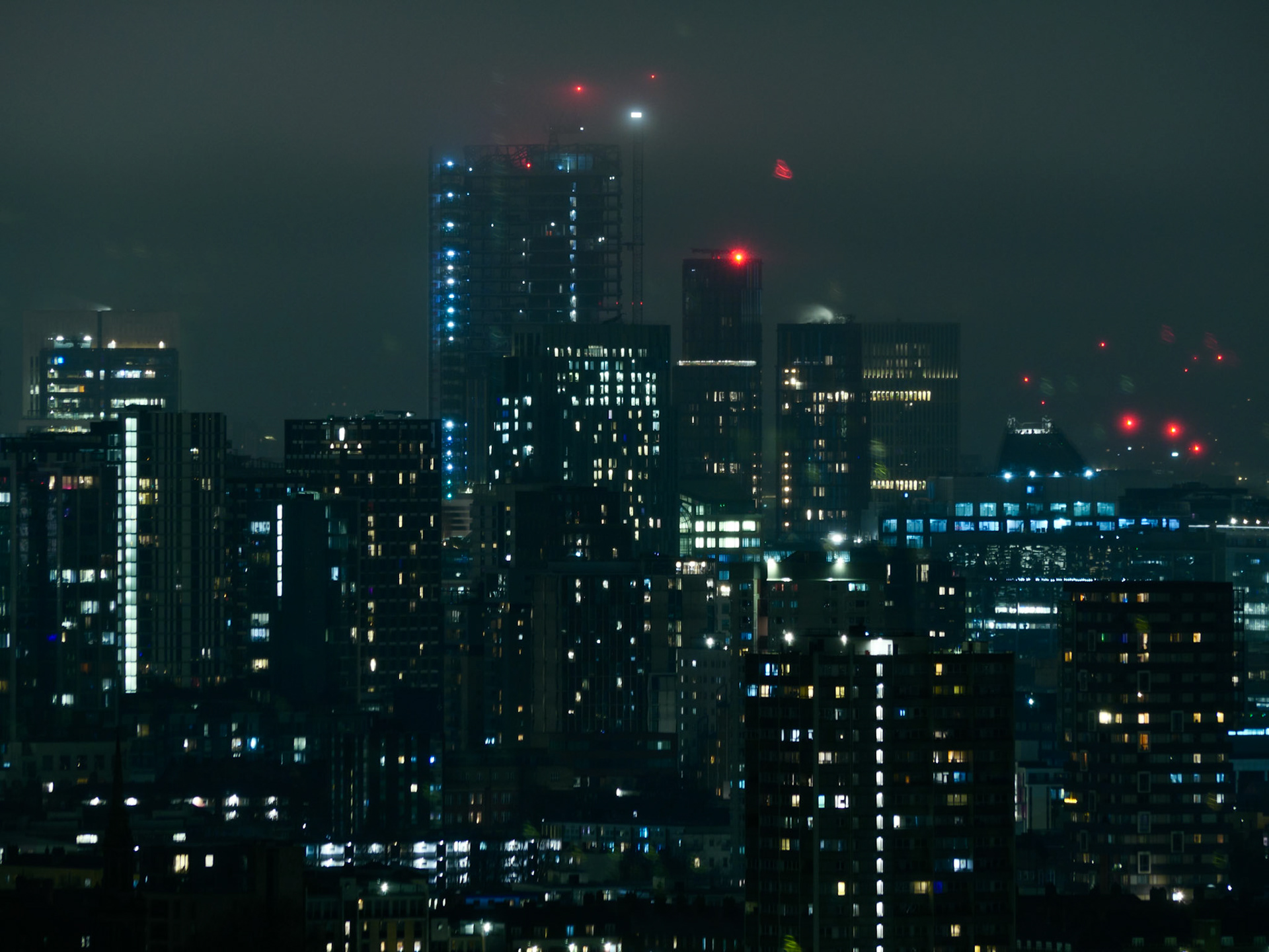 A moody, cyberpunk-style night cityscape of London featuring high-rise buildings and a skyscraper under construction glowing with neon blue lights, set against a dark, misty sky with red aviation warning beacons.
