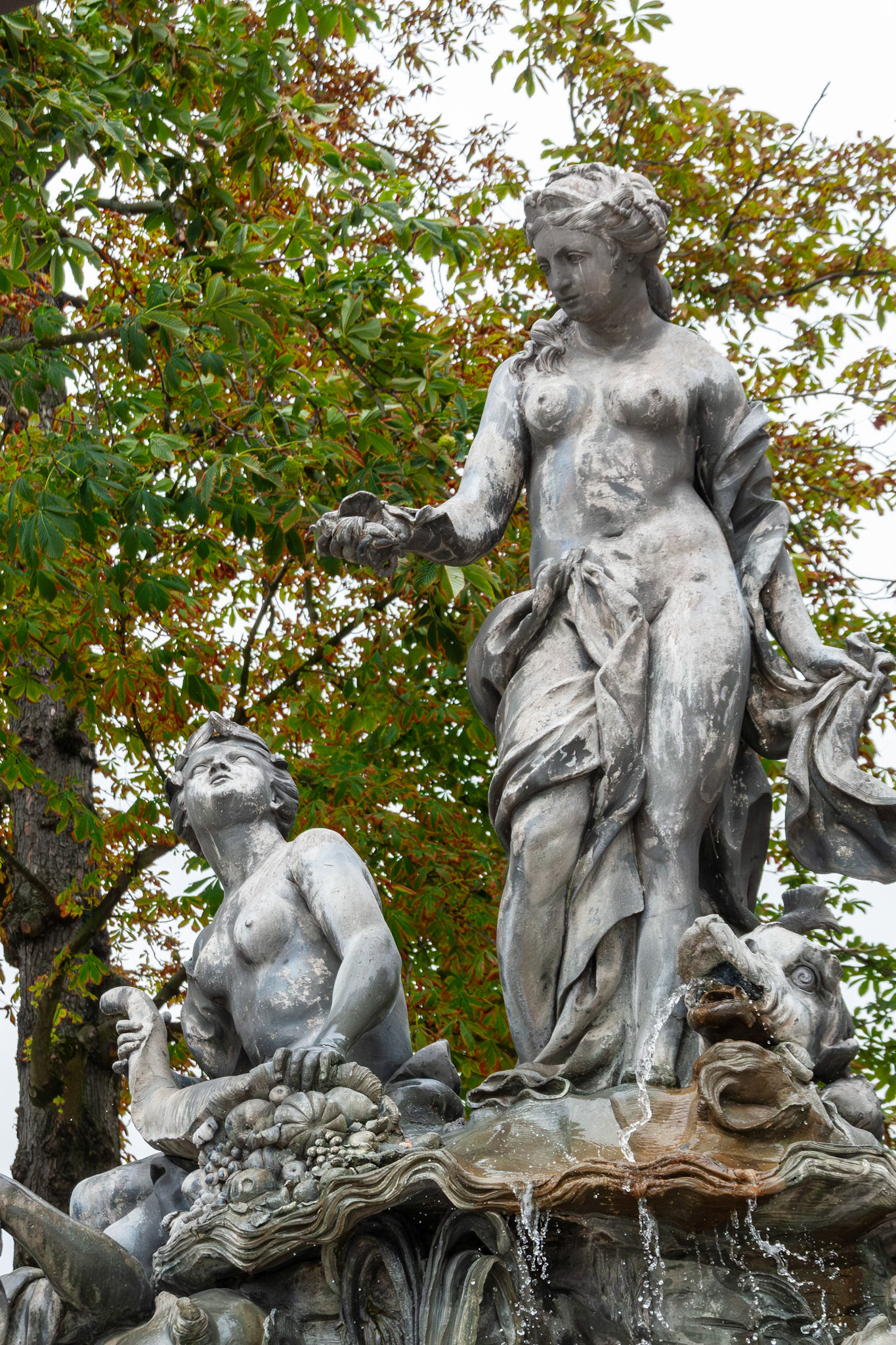 A detailed view of the Fountain of Neptune in Place Stanislas, Nancy, France, highlighting the elegant lead sculptures of Amphitrite and a sea nymph positioned against a backdrop of lush green chestnut trees.
