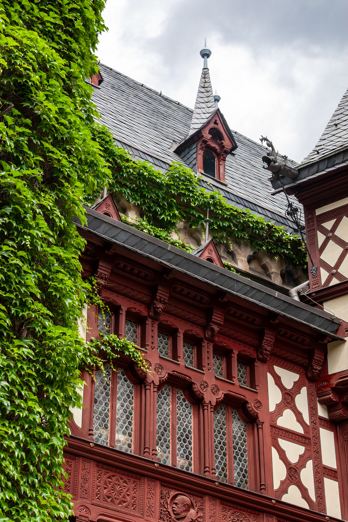 A detailed architectural shot of Wernigerode Castle in Germany, highlighting a vibrant red timber-framed facade with intricate carvings, leaded glass windows, and green ivy climbing the stone walls under a grey sky.