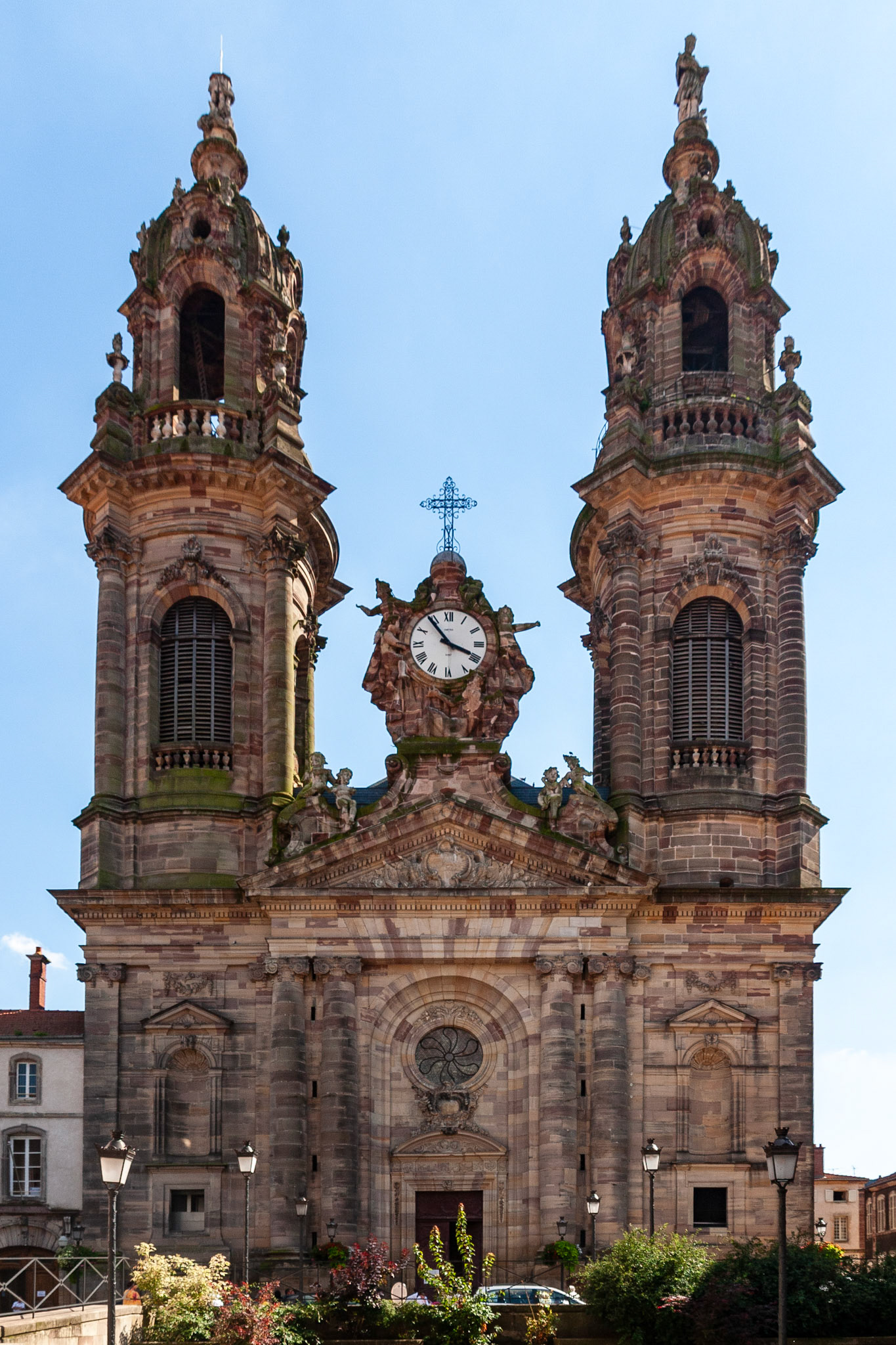 A full vertical view of a grand historic church in a French town, showcasing ornate Baroque architecture, symmetrical twin bell towers, a central clock, and a rose window under a clear blue sky.