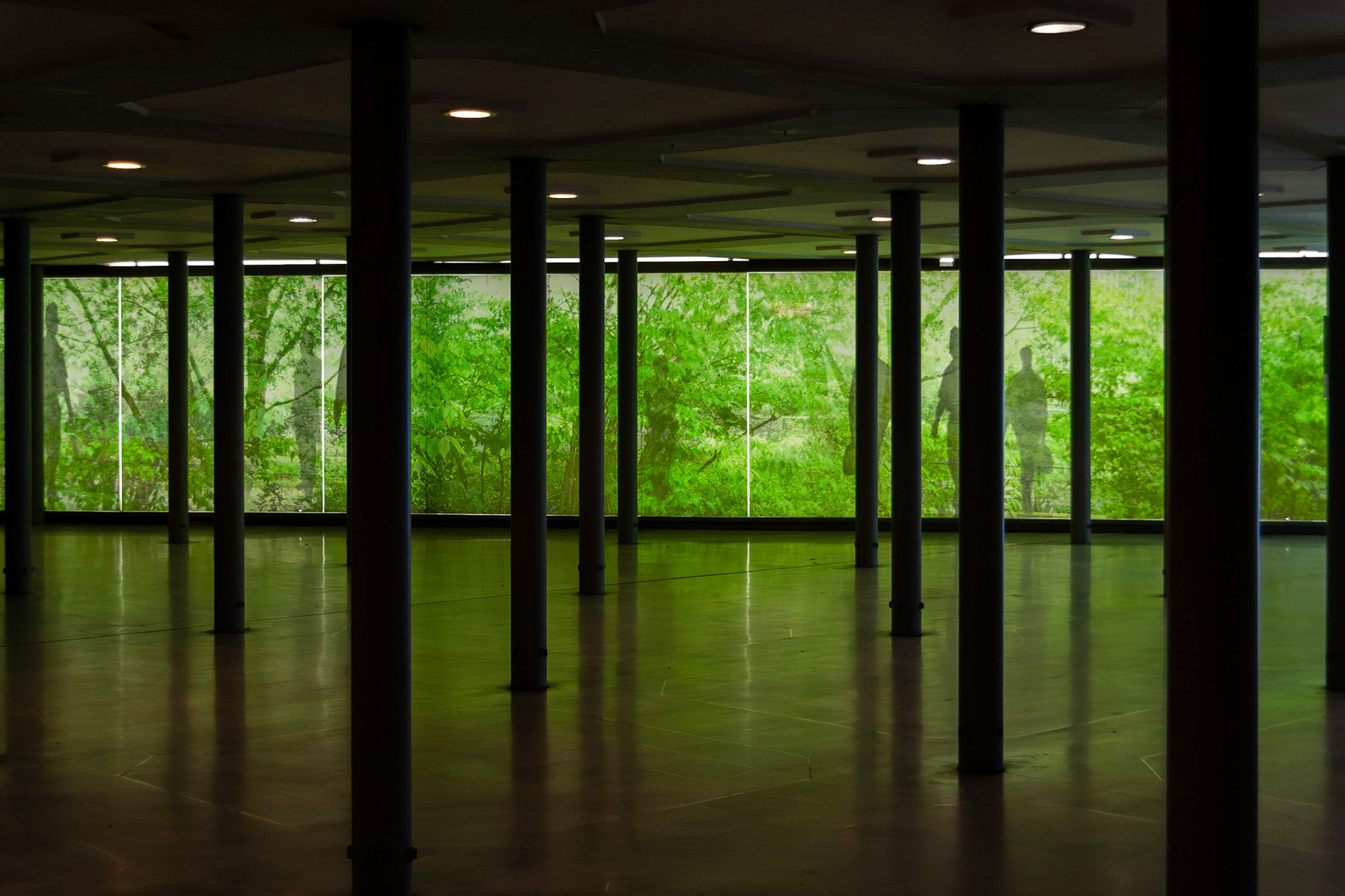 A contemporary interior space viewed from the shadows, featuring a series of dark vertical pillars that create a rhythmic perspective. A large panoramic window in the background reveals a lush green garden or forest, with faint silhouettes of people visible through the glass, blending nature with minimalist architecture.