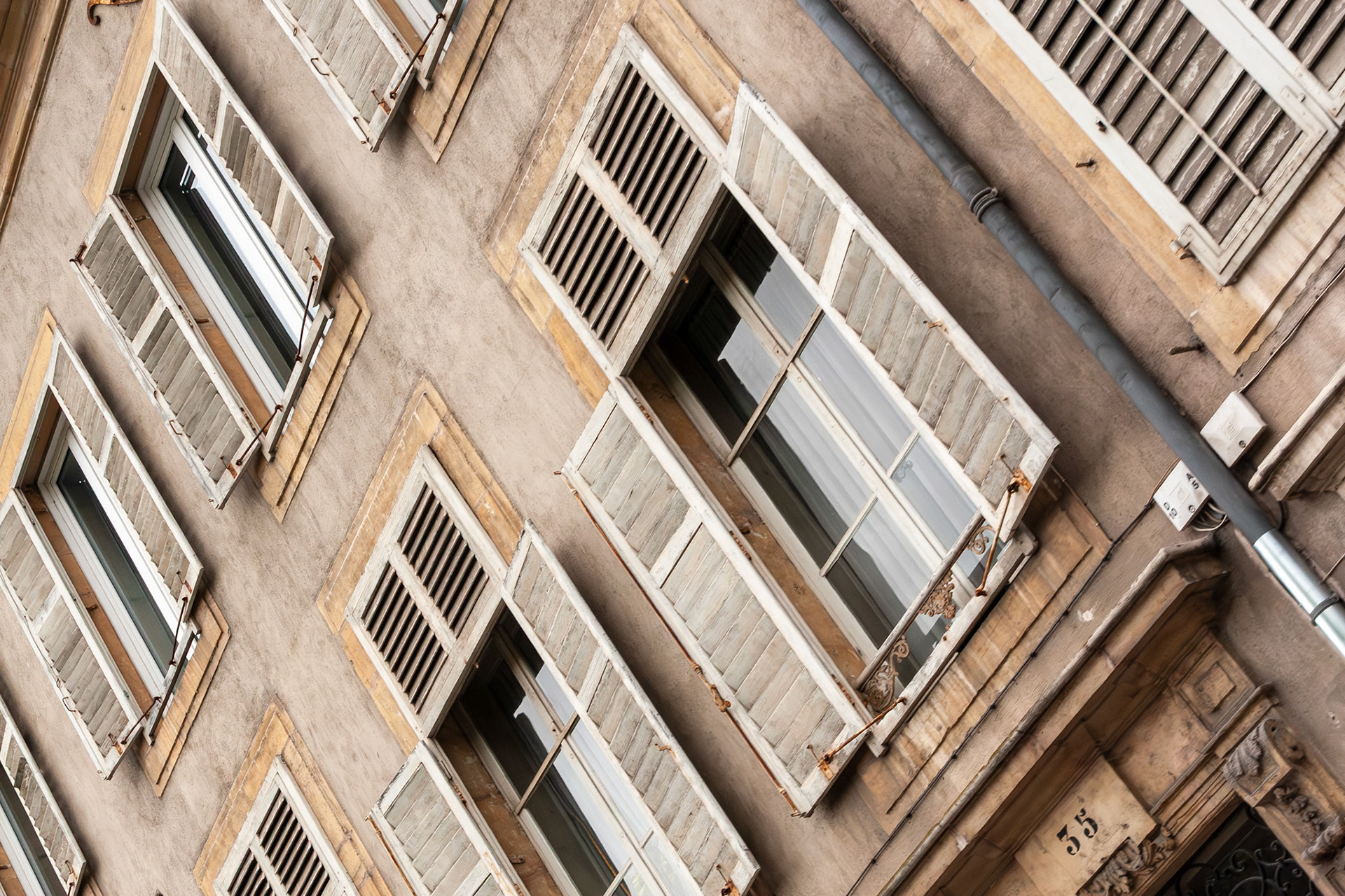 An angled architectural shot of a historic building facade in Nancy, France, featuring several windows with weathered grey wooden shutters and classic stone masonry.