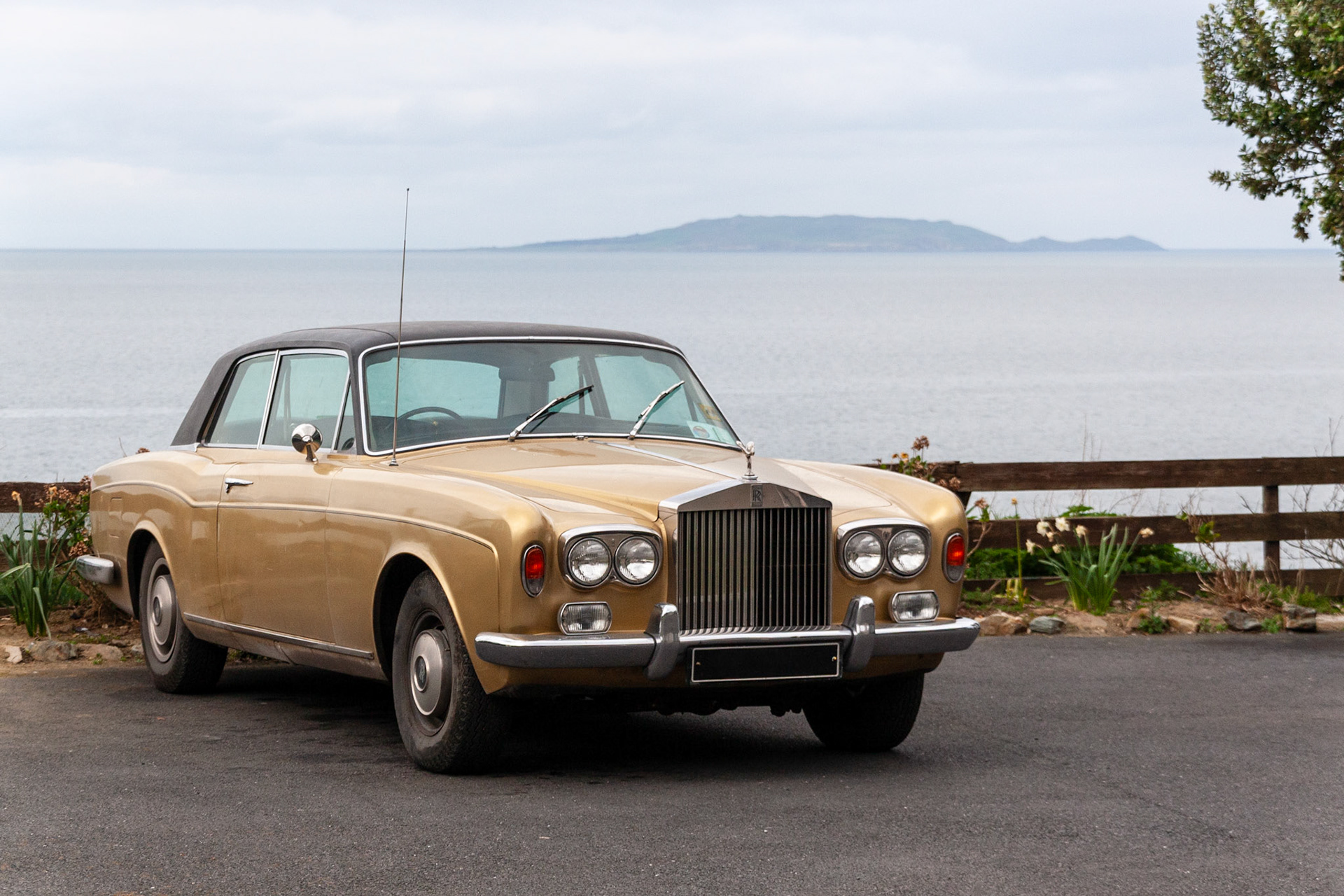 A luxurious gold vintage Rolls-Royce Corniche coupe parked along a scenic coastal road in Howth, Dublin, with the Irish Sea and Ireland's Eye island visible in the background.