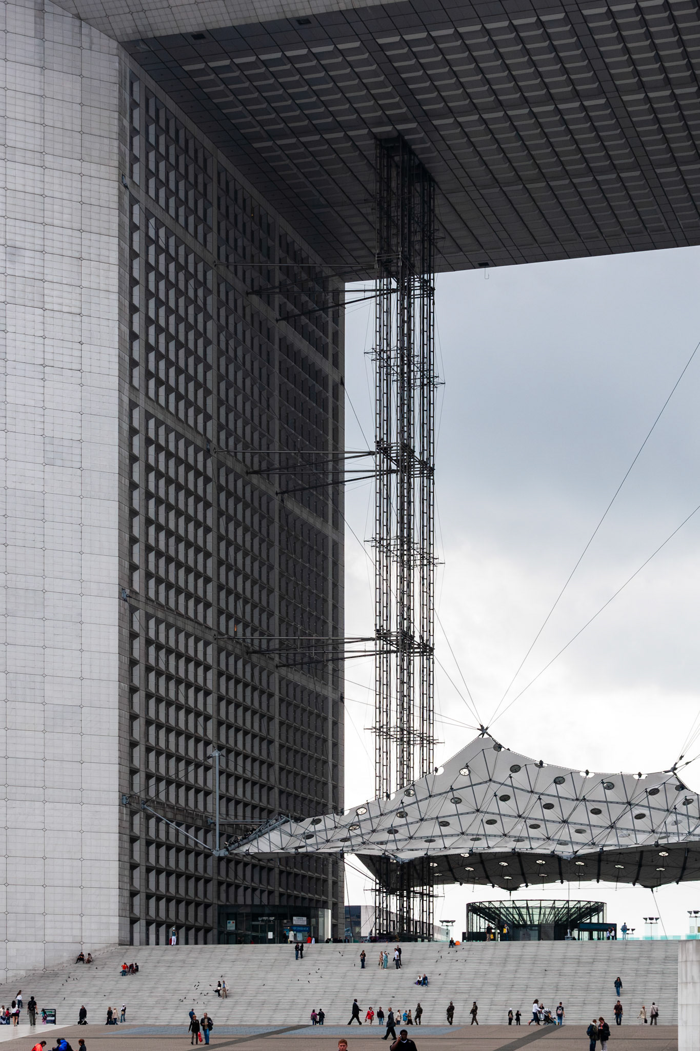 A vertical low-angle shot of a massive, cube-shaped modern building featuring a repetitive grid facade. The structure shelters a large public plaza with wide stairs and a unique white geometric tensile canopy. Pedestrians provide a sense of scale against the towering grey and white architectural elements under a soft, overcast sky.