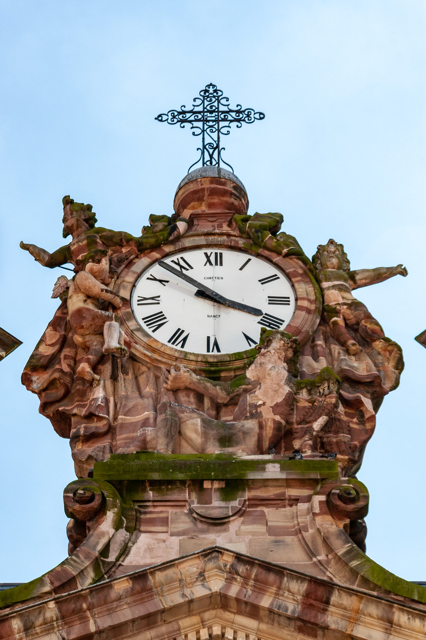 A detailed low-angle view of a historic stone church clock in France, featuring intricate Baroque carvings of cherubs and figures, topped with a decorative iron cross against a clear blue sky.