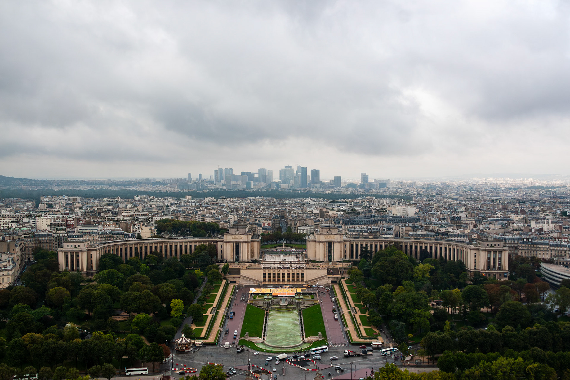 A high-angle wide shot of the Palais de Chaillot and the Trocadero Gardens in Paris, France. The image captures the symmetrical architecture of the museum complex, the central fountain, and the surrounding cityscape stretching toward the La Defense business district on a moody, cloudy day.