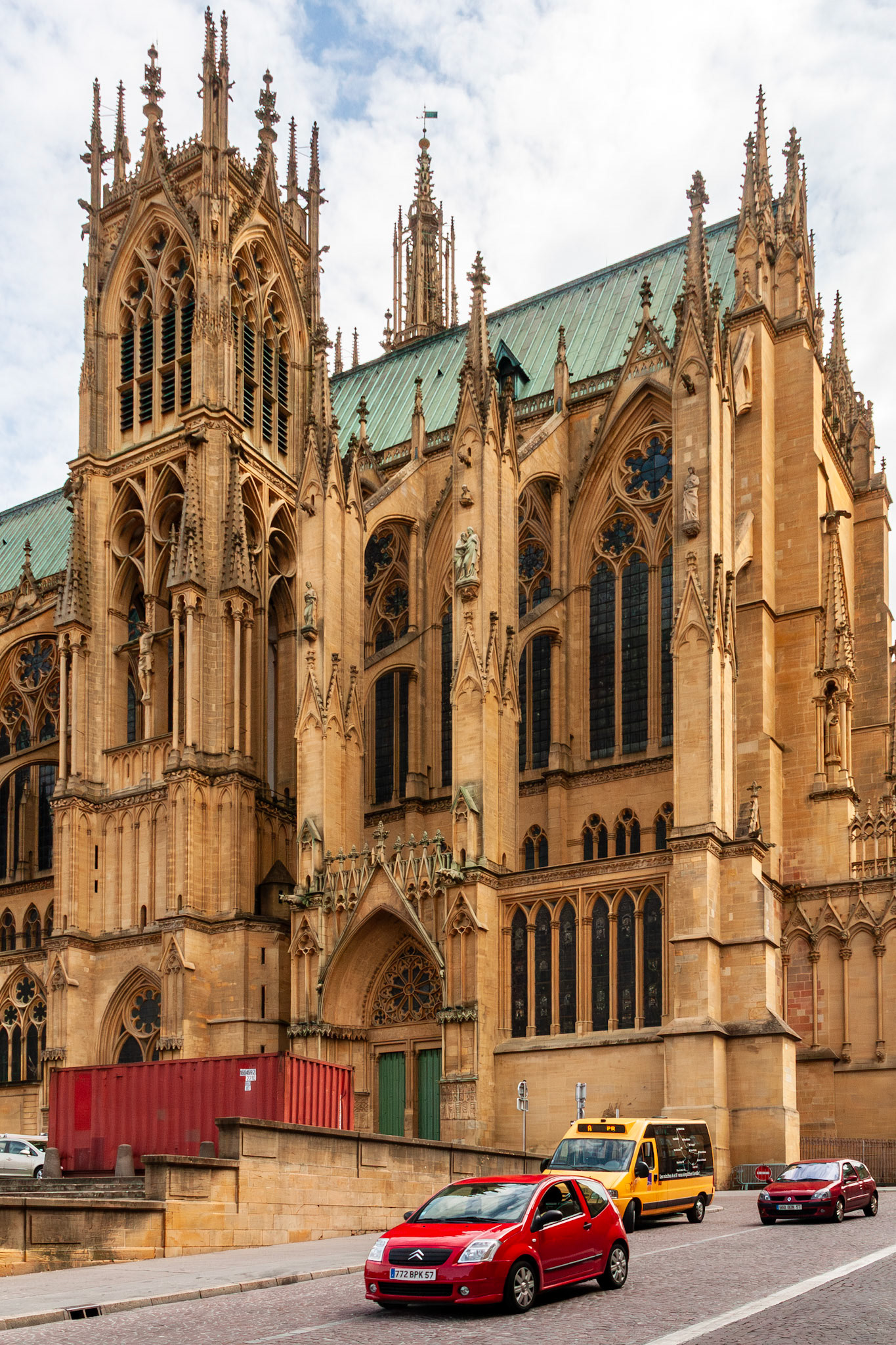 A majestic view of the Cathédrale Saint-Étienne (St. Stephen's Cathedral) in Metz, France, showcasing its soaring yellow limestone Gothic architecture, intricate flying buttresses, and ornate spires as cars pass by on the cobblestone street below.