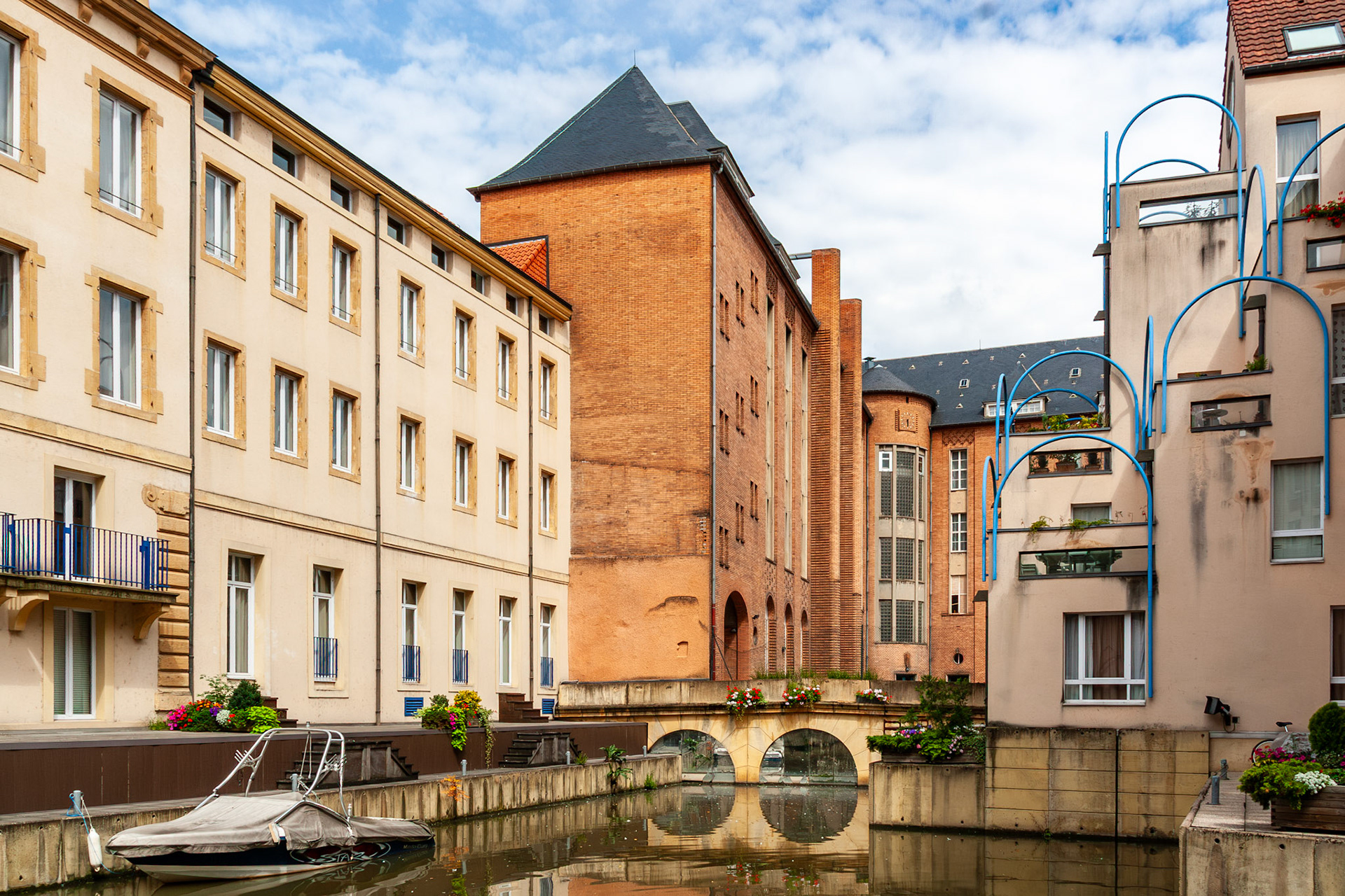 A contemporary urban scene in Metz, France, featuring multi-story residential buildings with unique architectural details like blue arched railings and brick facades along a quiet city canal. A small motorboat is docked in the foreground, reflecting the blend of modern living and historic European waterways.
