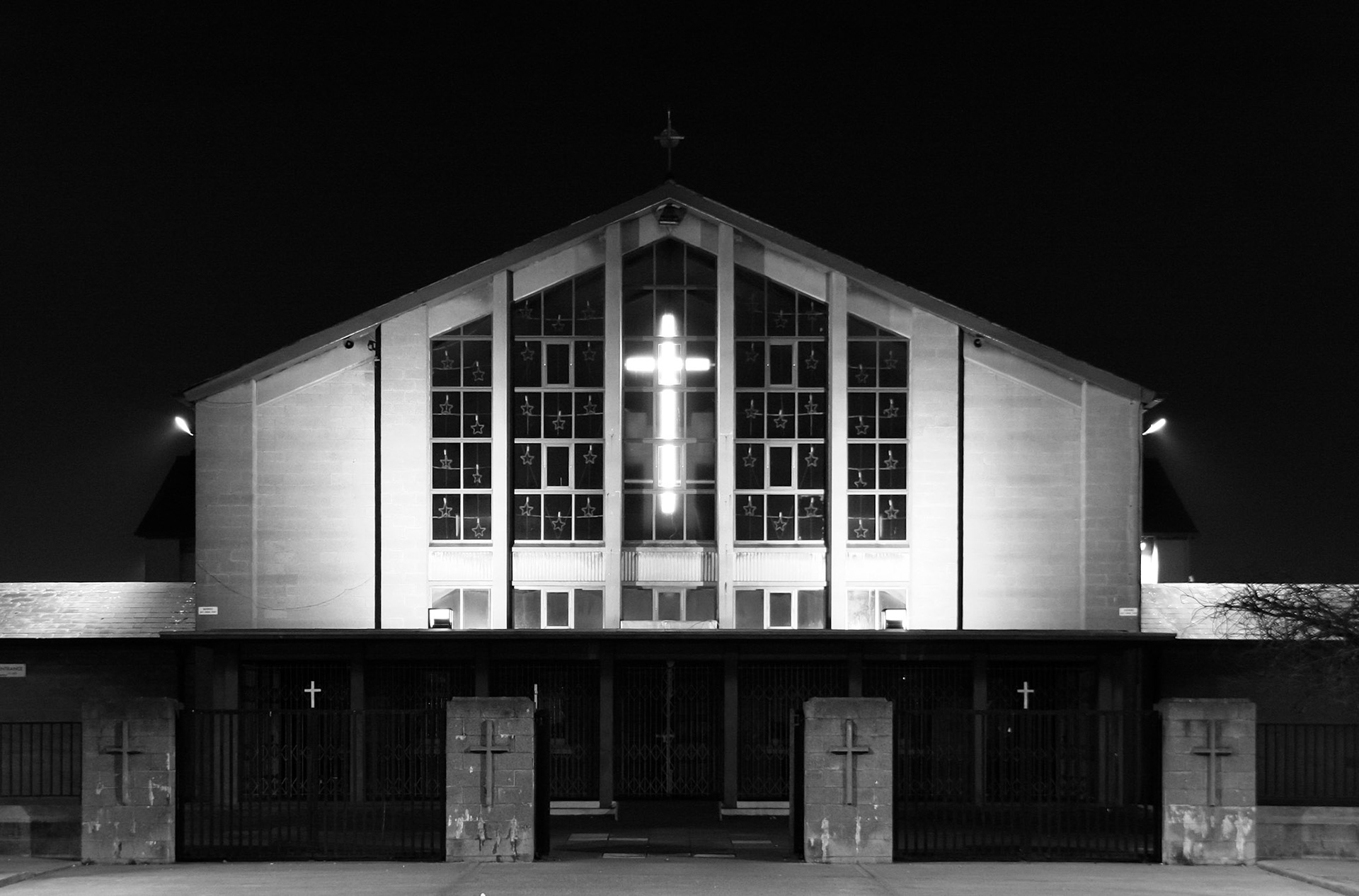 A striking black and white long-exposure photograph of the Church of Our Lady of the Assumption in Ballyfermot, Dublin. The image features a symmetrical architectural composition at night, highlighting the glowing illuminated cross on the glass facade and the dark, moody sky over the suburban Irish landmark.