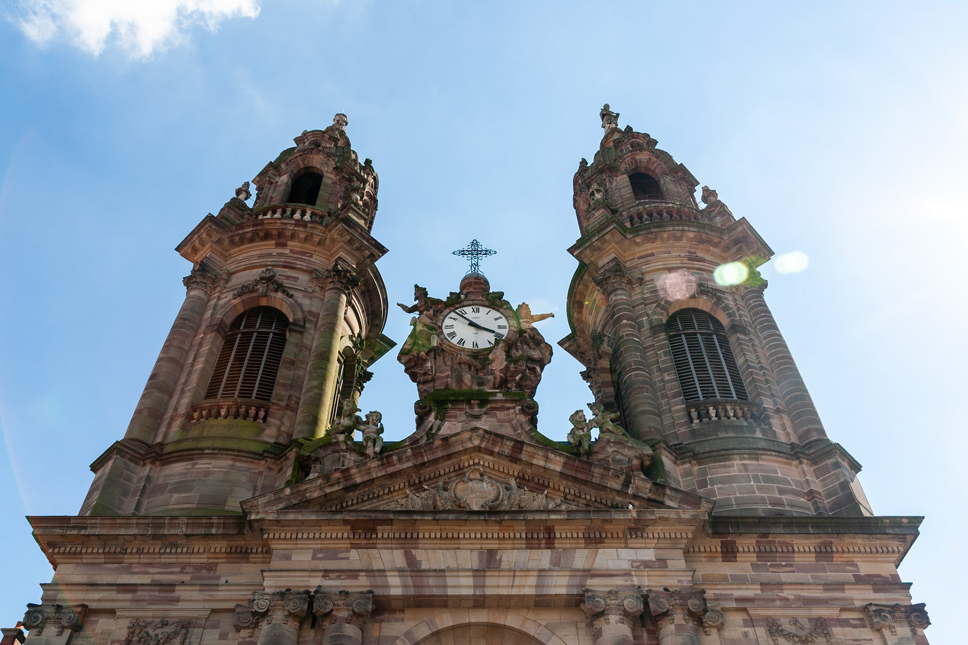A low-angle architectural shot of a grand French church featuring symmetrical twin bell towers, intricate Baroque stonework, and a central ornate clock under a bright blue sky with subtle lens flare.
