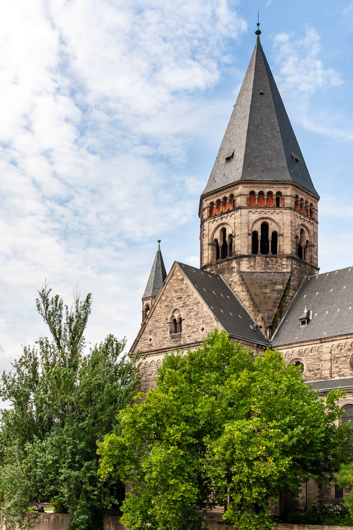 A bright, daylight view of the Temple Neuf, a grey sandstone Protestant church built in the Neo-Romanesque style, partially framed by lush green trees against a scattered cloud sky in Metz, France.