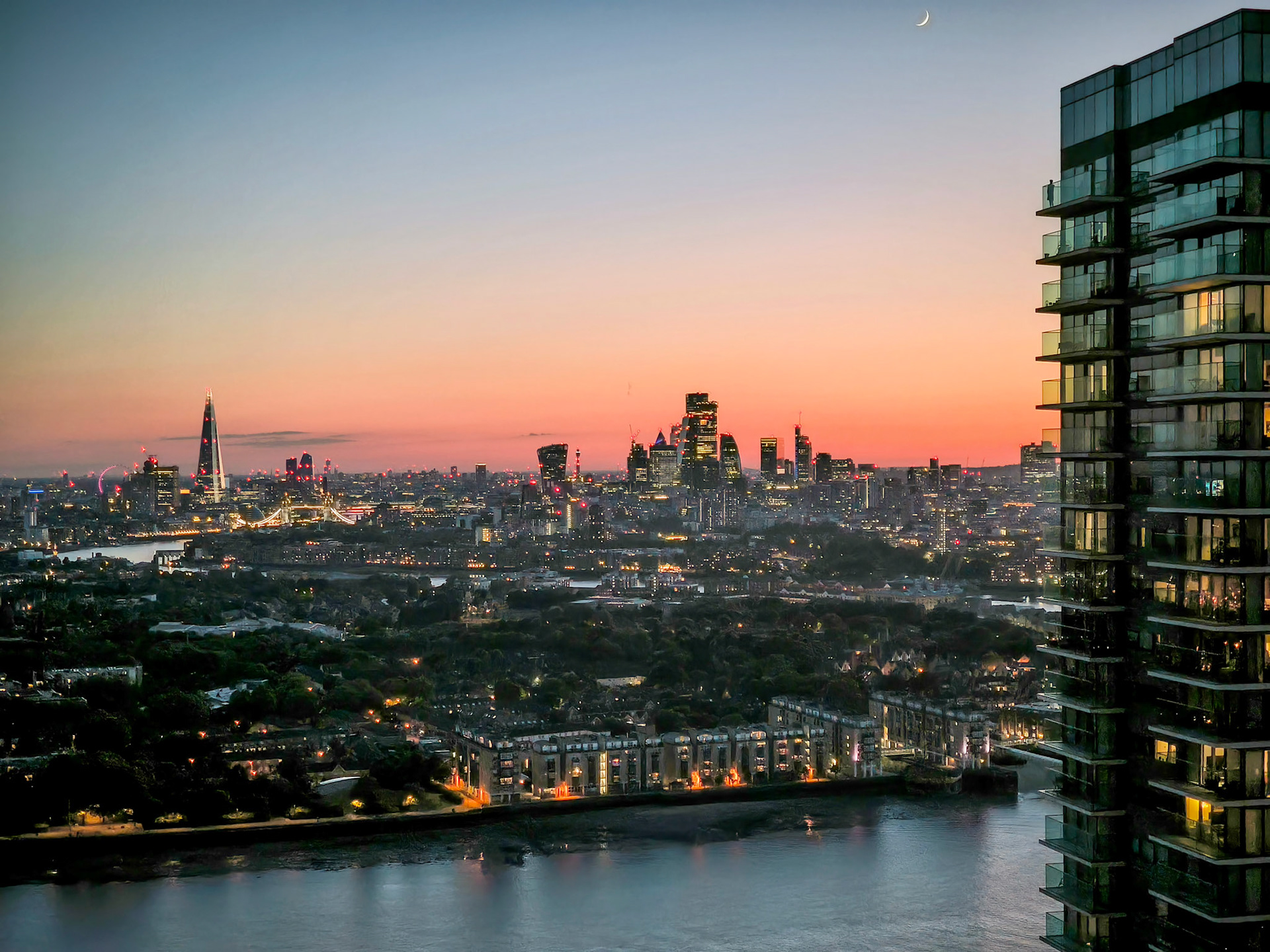 A breathtaking panoramic view of London at dusk, featuring iconic landmarks like The Shard and Tower Bridge under a vibrant orange sky with a crescent moon overhead.