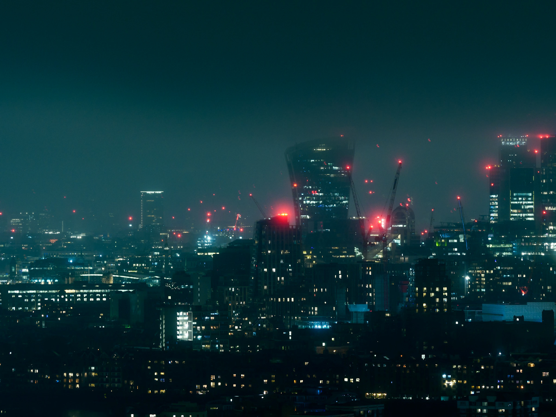 A high-angle, atmospheric night view of the London cityscape featuring illuminated skyscrapers, construction cranes, and glowing red aviation lights under a misty, dark sky, capturing a moody cyberpunk-inspired urban environment.