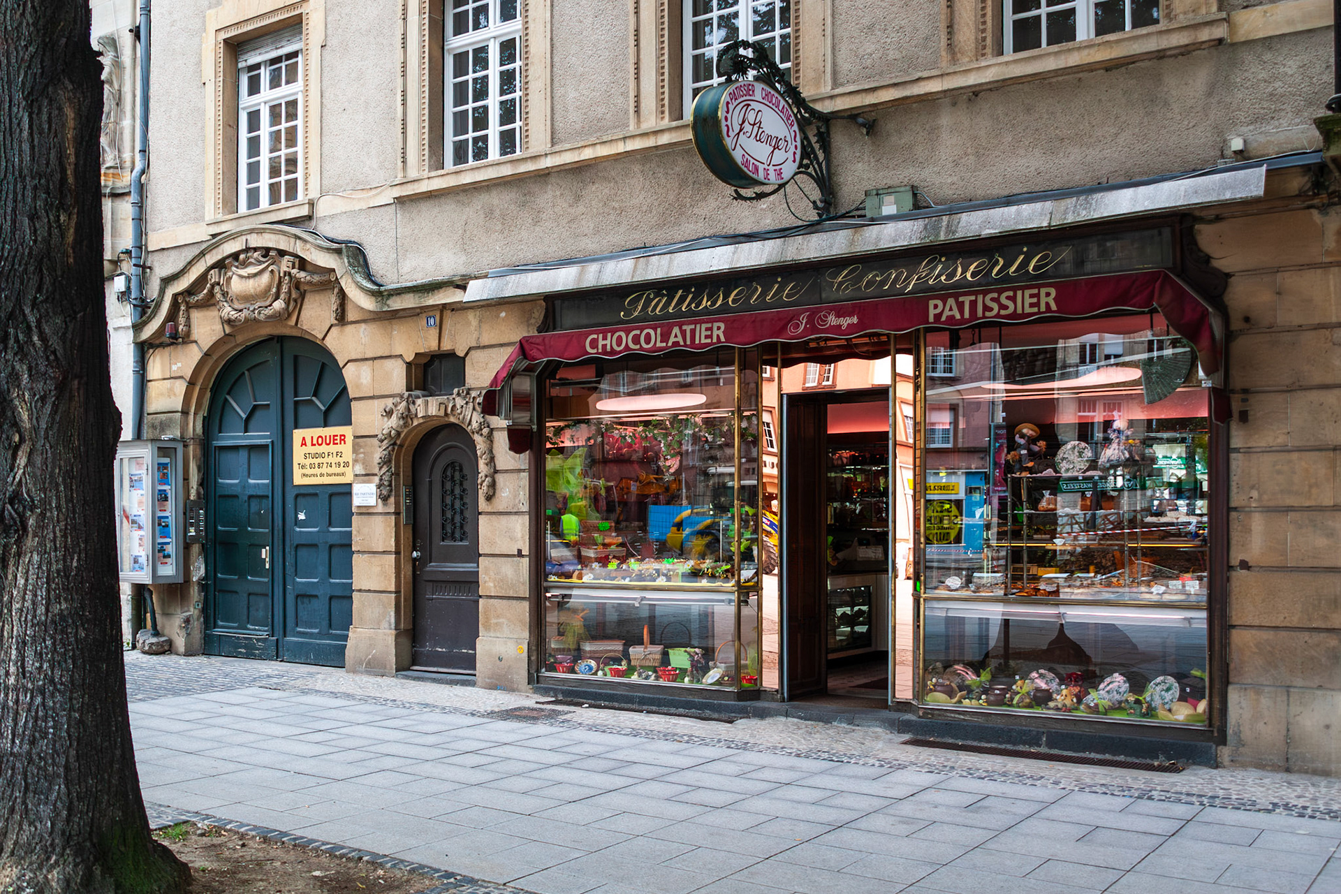 The charming exterior of J. Stenger, a traditional French pâtisserie and chocolaterie in Metz, France. The shop features a classic red awning, large glass display windows filled with artisanal sweets, and sits next to a historic arched stone doorway.