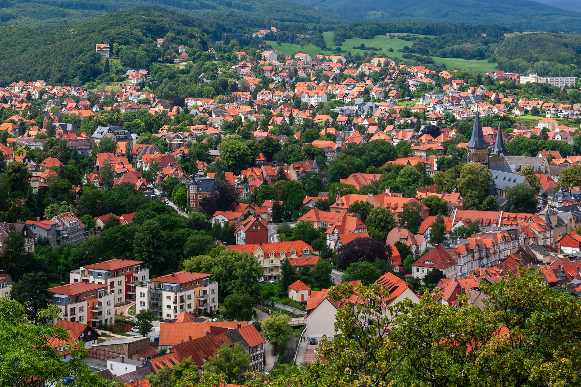 A wide-angle elevated view of the picturesque town of Wernigerode, Germany, showcasing a dense tapestry of traditional red-tiled roofs, historic church spires, and lush green forests extending into the Harz mountains.
